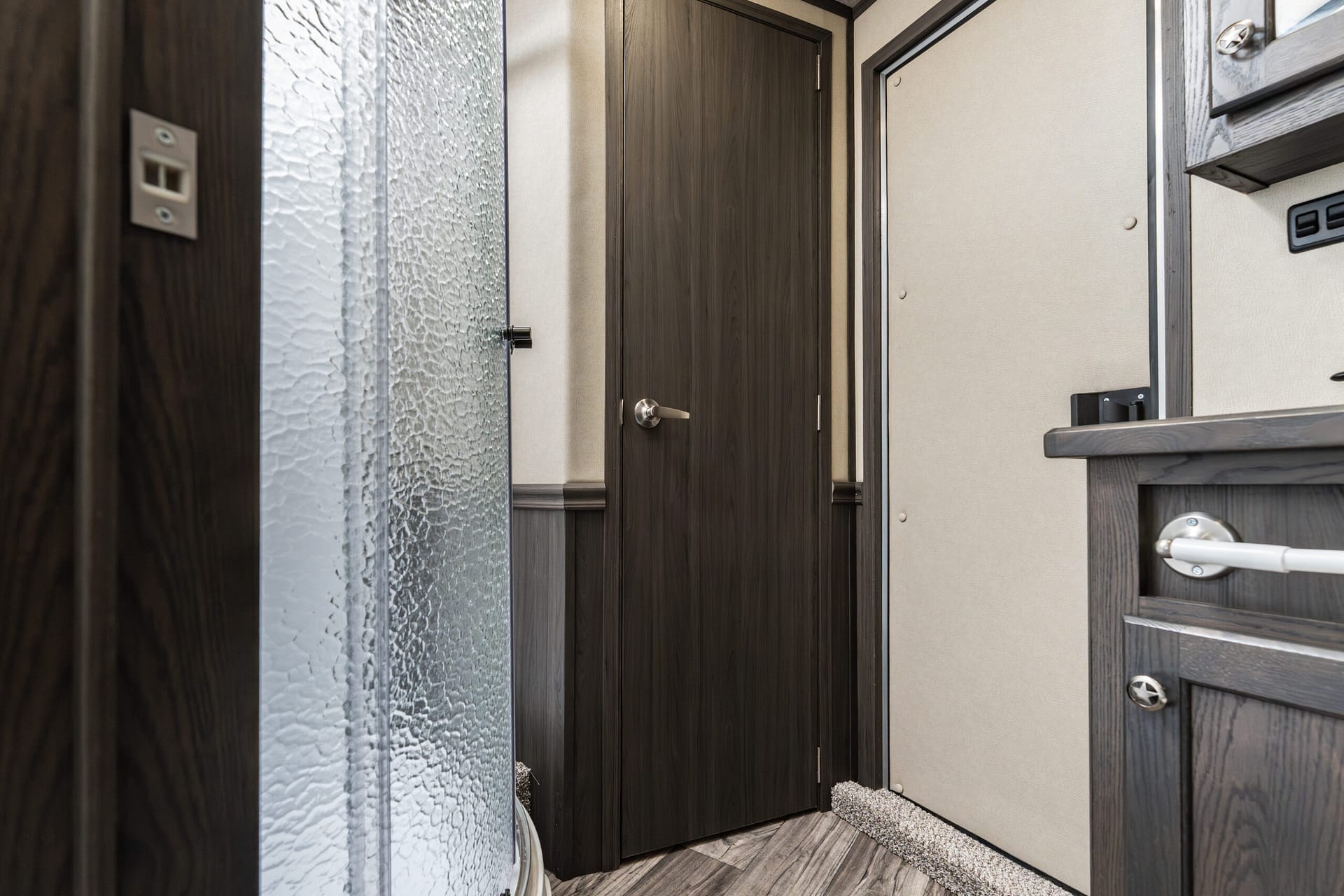 Narrow interior hallway with dark wood doors, silver handles, textured glass panel on the left, beige walls, and a small shag rug on the floor.