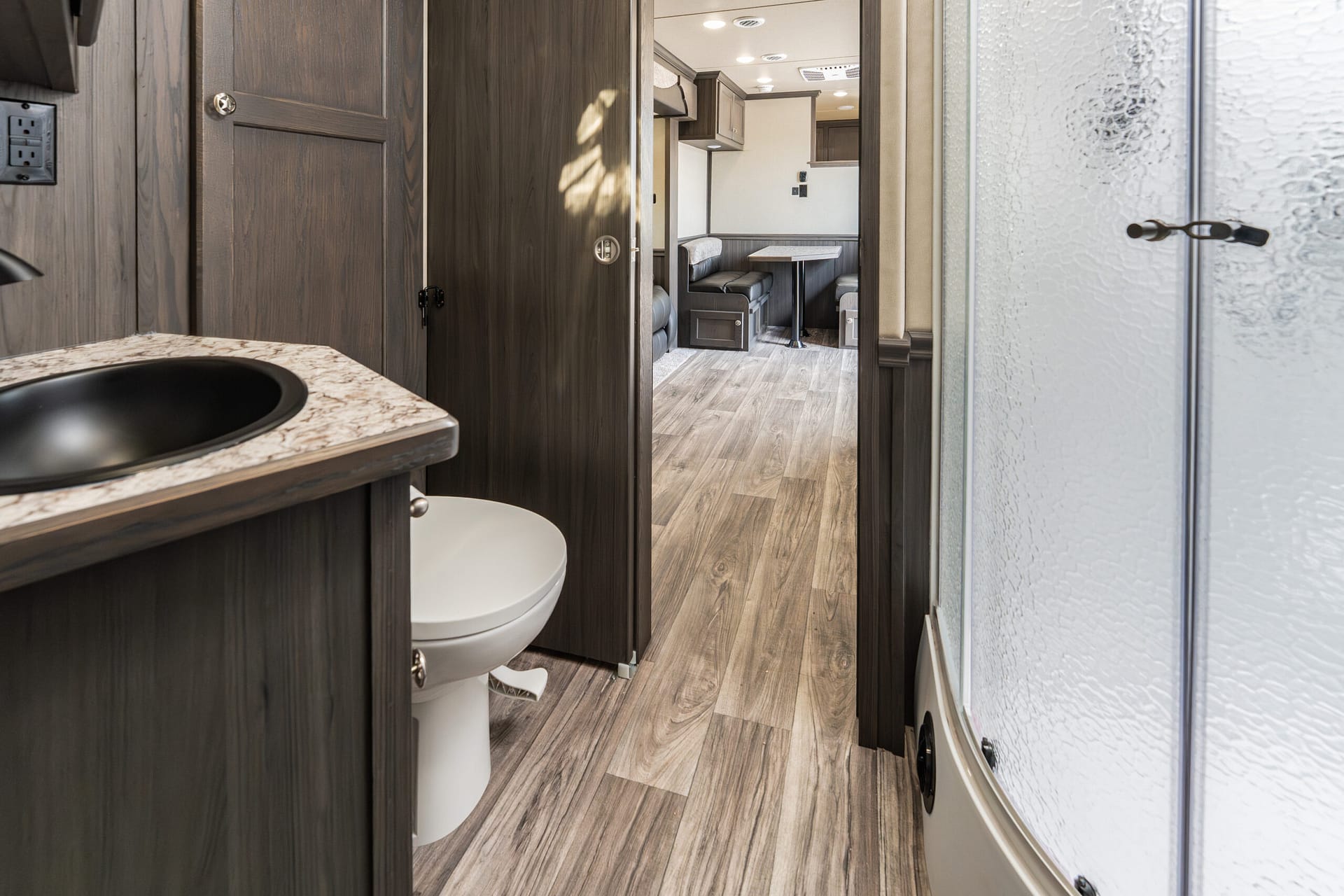 Interior of an RV bathroom with a white toilet, dark wood cabinets, and a marble countertop, open to a small dining lounge area in the background.