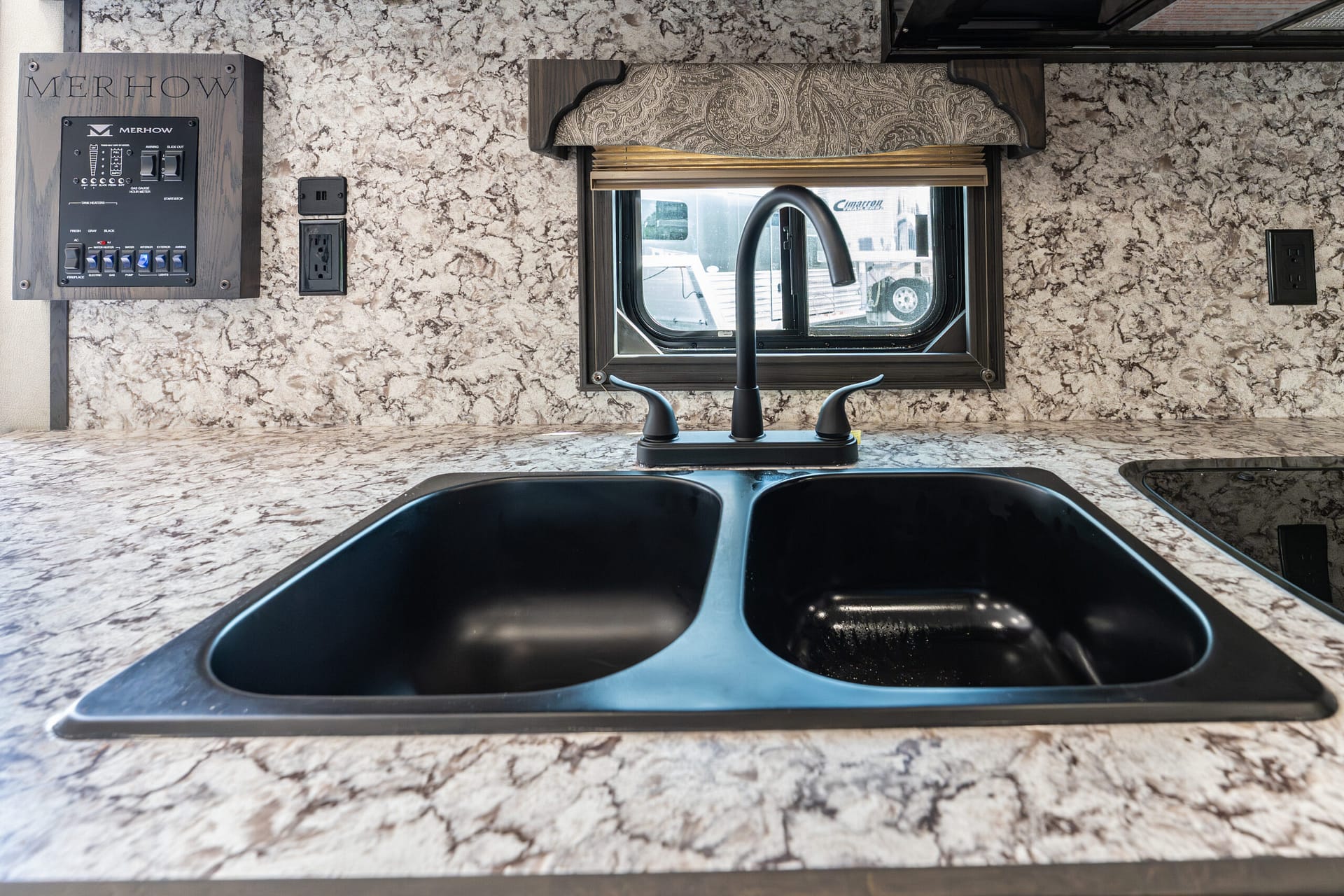 RV kitchen sink area with a black double basin sink set in a beige marble countertop, black gooseneck faucet, and a window with a decorative valance above; panel and outlets on the left show controls and power.