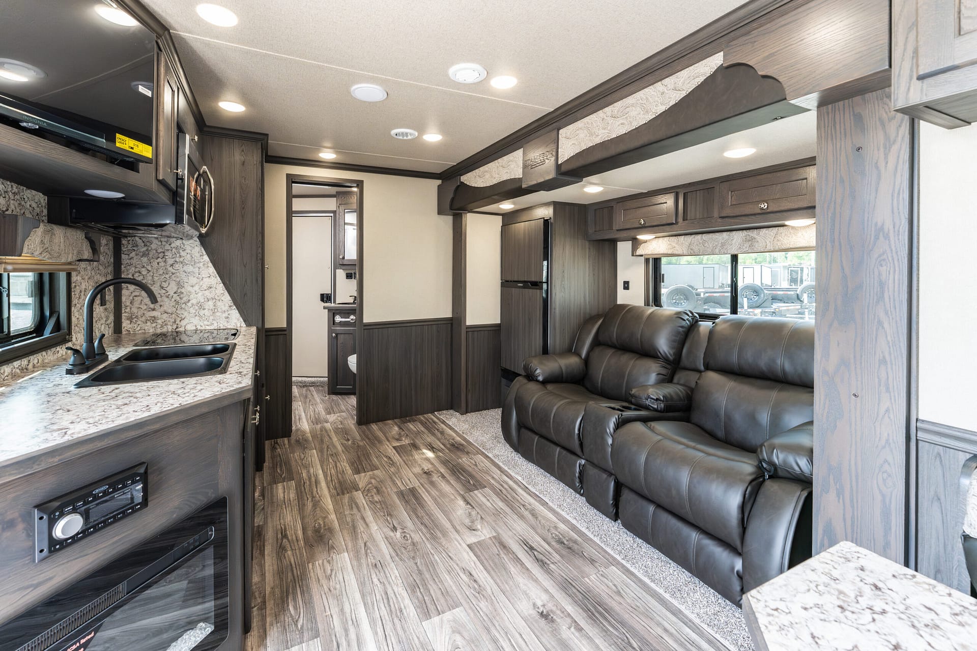 Interior of an RV kitchen and living area with dark wood cabinetry, granite countertops, a double sink, and a black leather sofa by the windows.