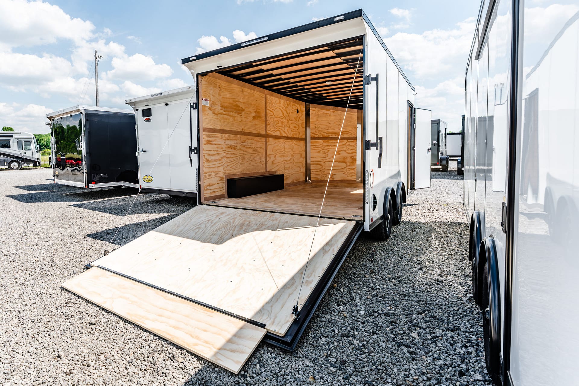 Open cargo trailer with ramp lowered, showing plywood walls and wooden floor in a gravel lot.