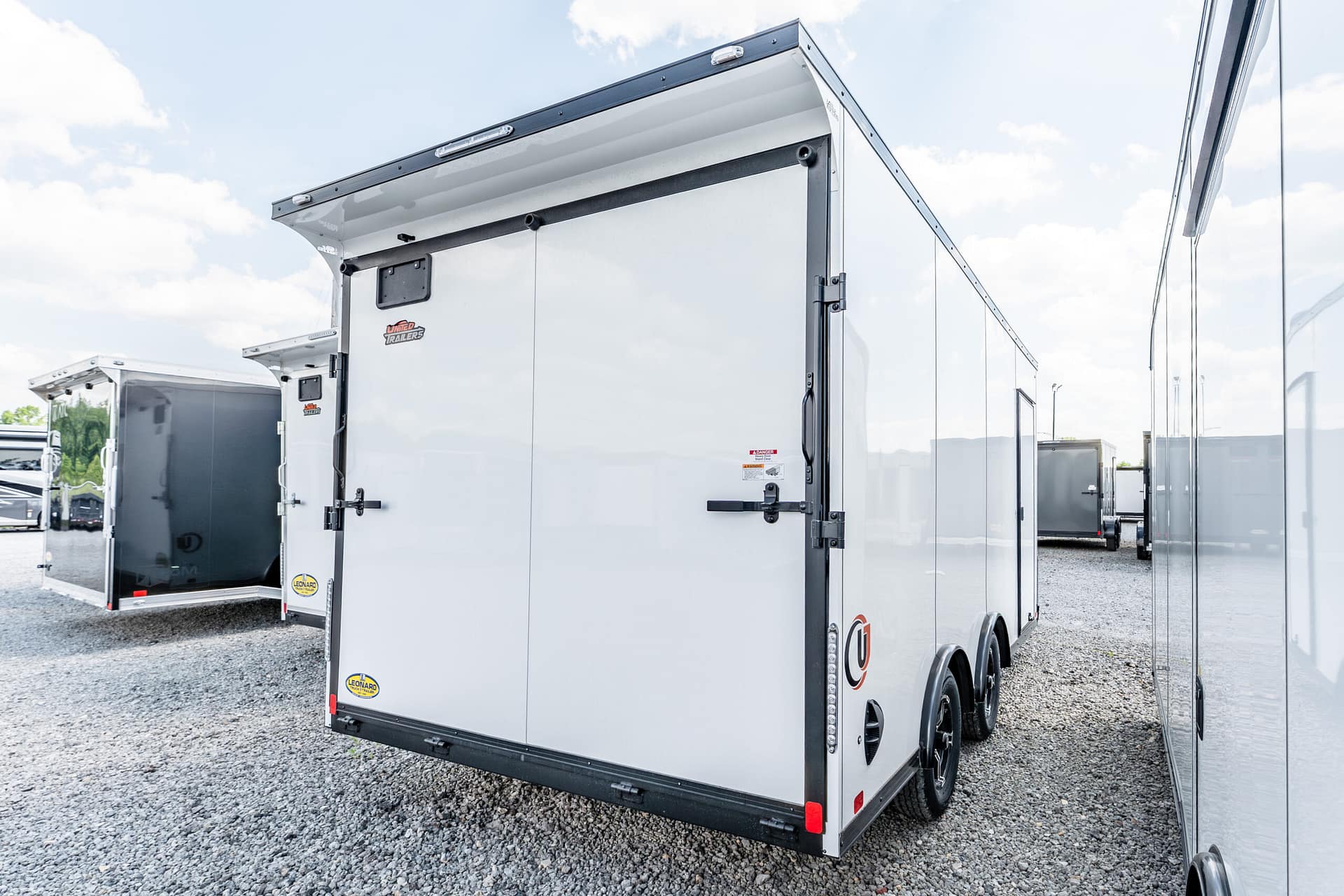 Side and rear view of a large silver enclosed trailer in a gravel lot, with another trailer visible in the background on a sunny day.
