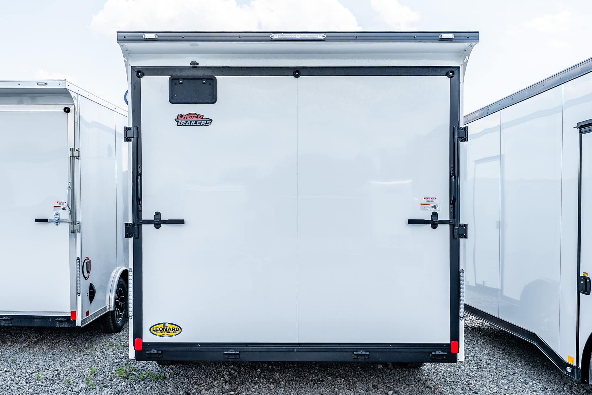 Rear view of a white enclosed trailer with double doors, black latches, and small branding sticker at the lower left corner over gravel ground.