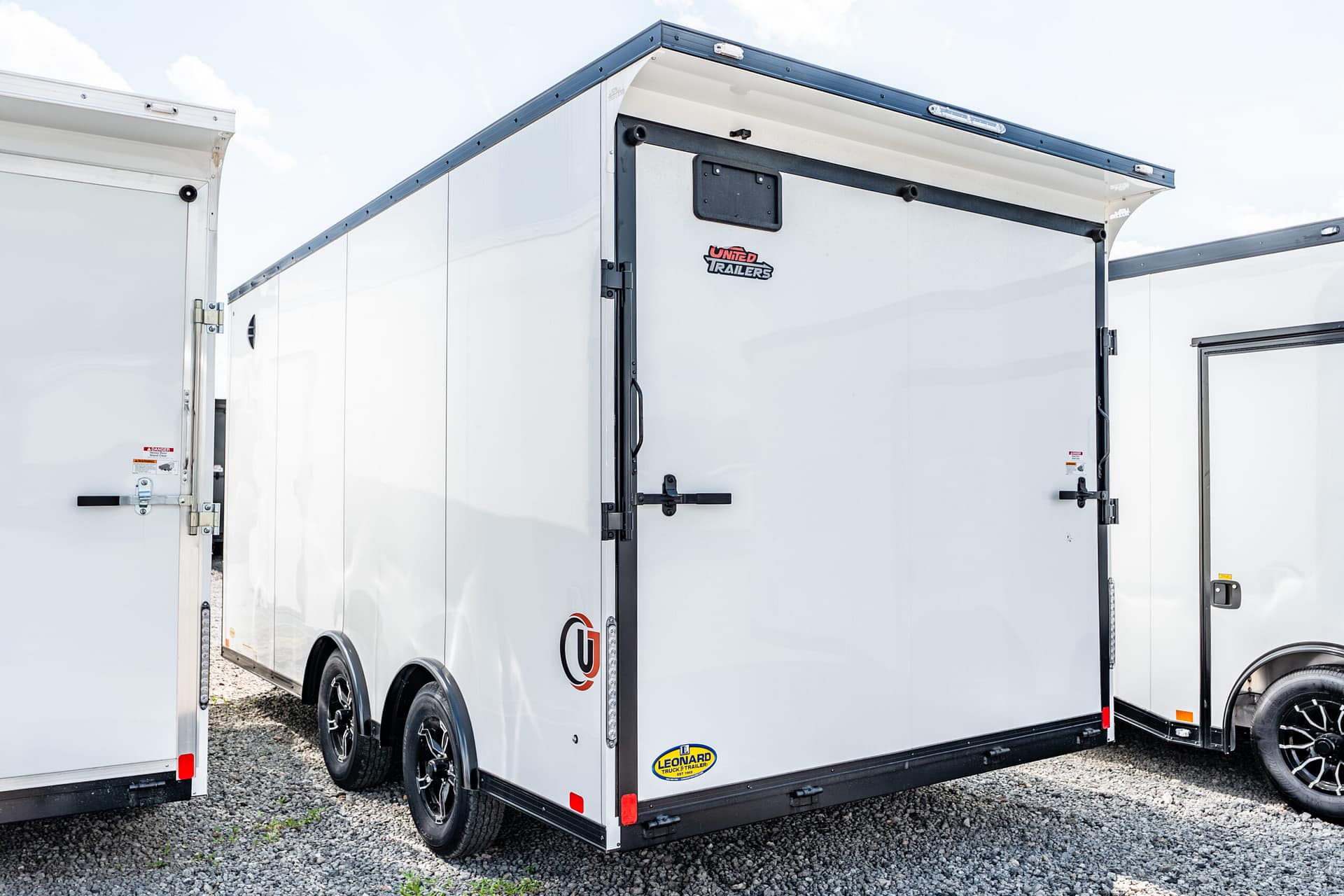 White tandem-axle enclosed cargo trailer with black trim and rear doors, parked on gravel, showing brand decals (Leonard, United Trailers).