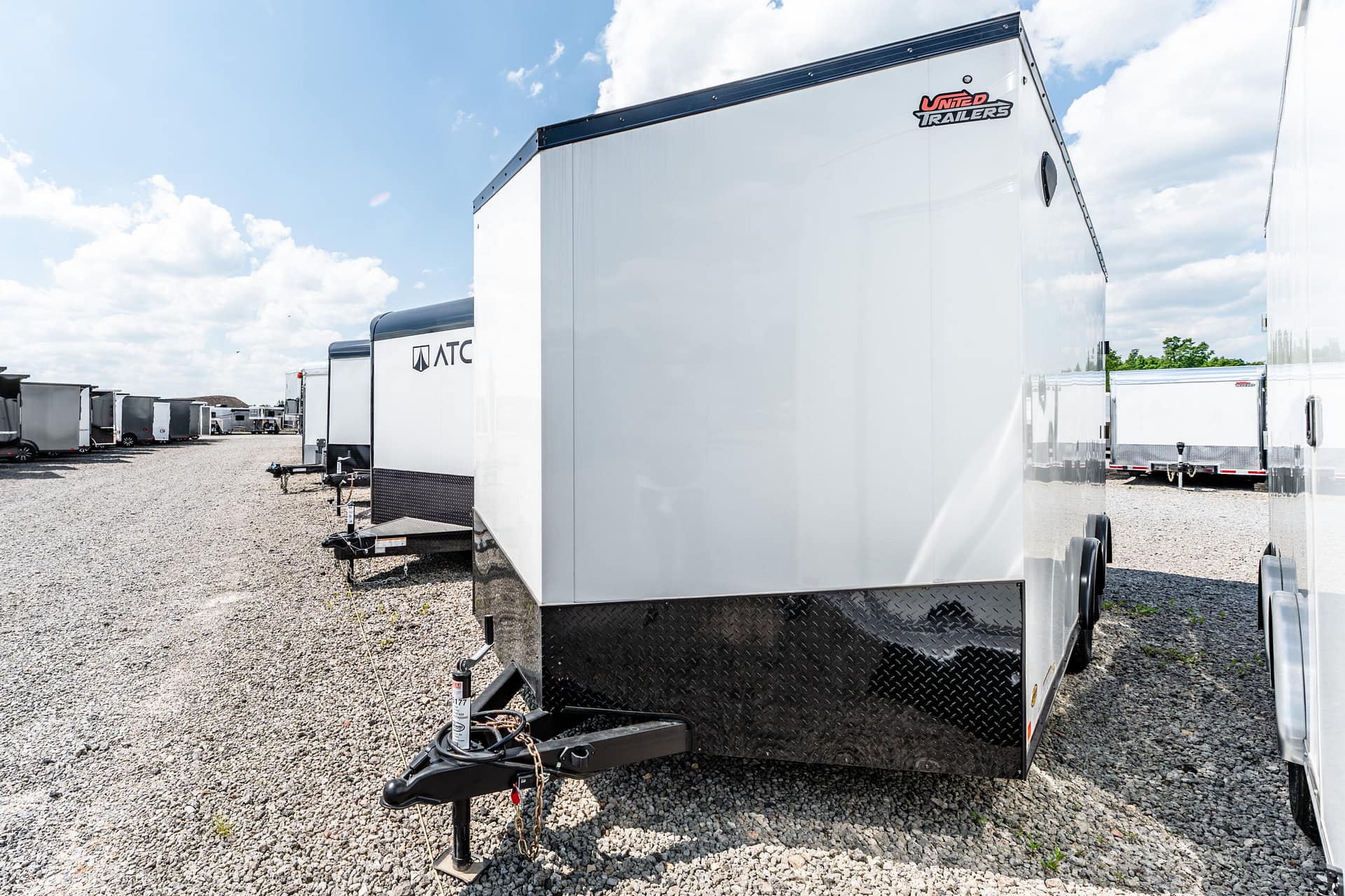 White enclosed cargo trailer in the foreground with a diamond‑plate front, hitch and chains visible, row of trailers on gravel under a blue sky.