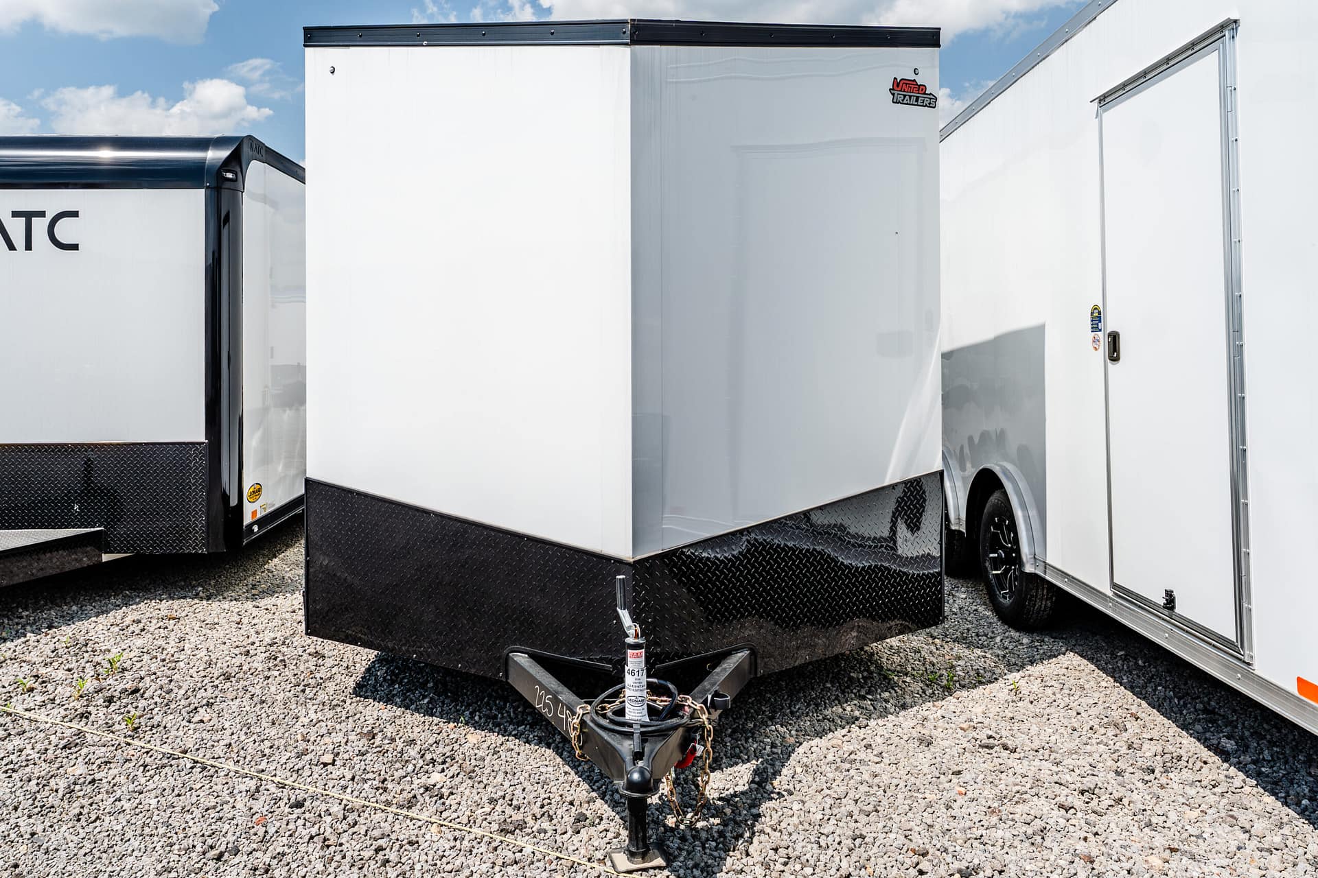 Close-up of a black-trimmed white cargo trailer hitch on a gravel lot, with other white trailers in the background and a diamond-plate kick panel at the bottom.