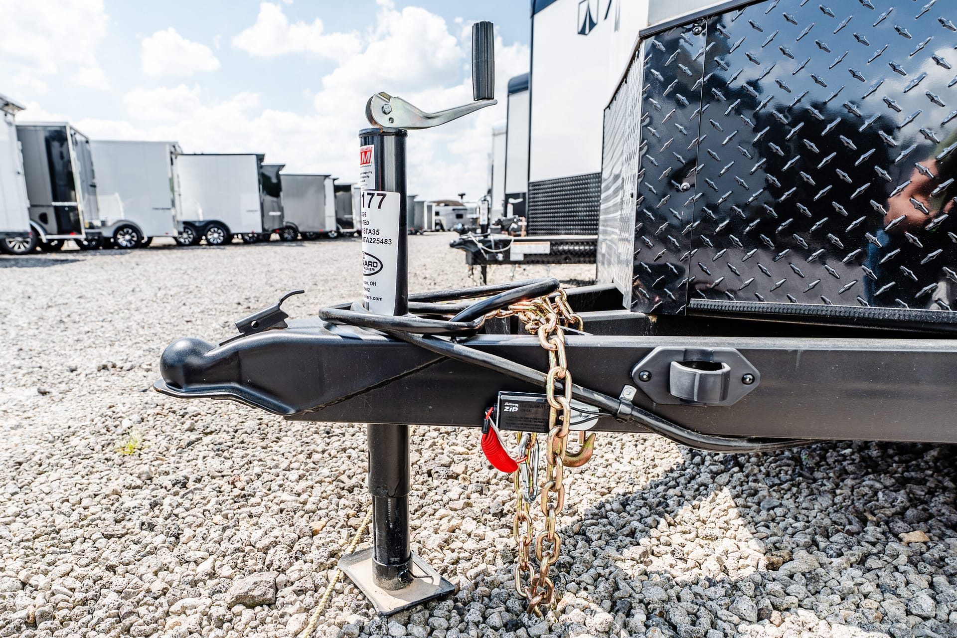 Close-up of a trailer hitch jack with chains on a gravel lot, with several enclosed trailers in the background.