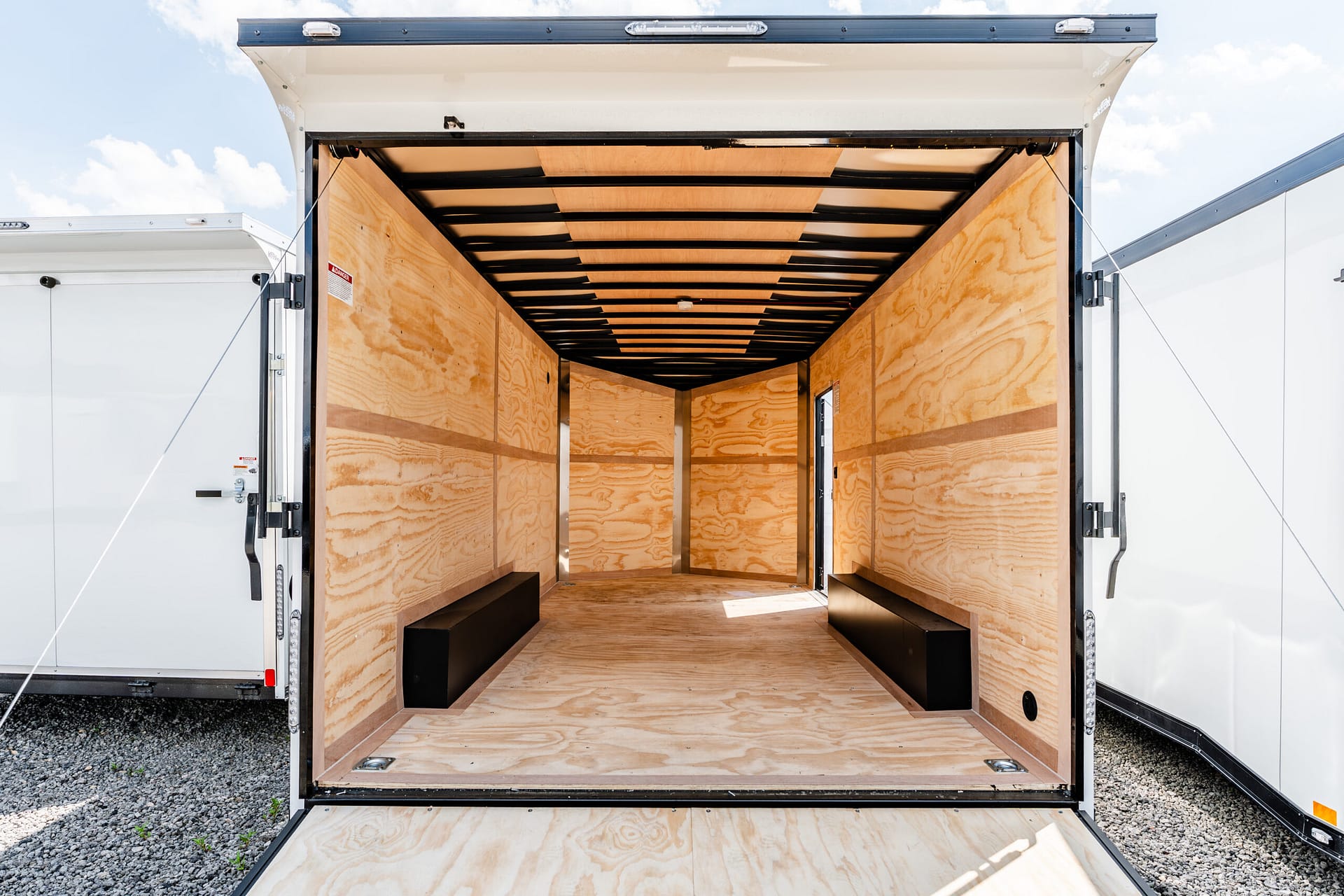Open rear of an empty wooden cargo trailer with plywood walls and floor, exposed ceiling beams, and daylight entering from outside to load items.