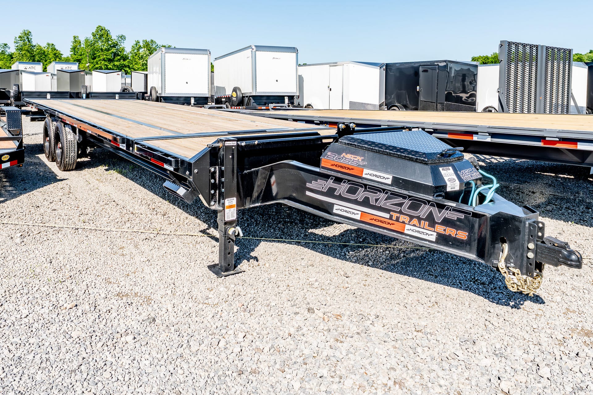 Flatbed trailer in a gravel lot, with multiple enclosed trailers and equipment in the background under a clear blue sky.