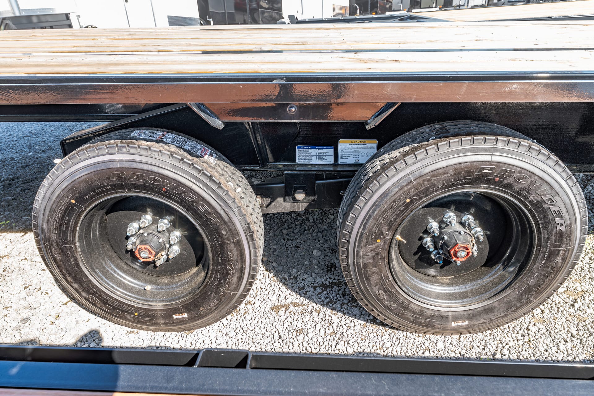 Dual-axle trailer wheel assembly with a wooden deck and gravel ground beneath.