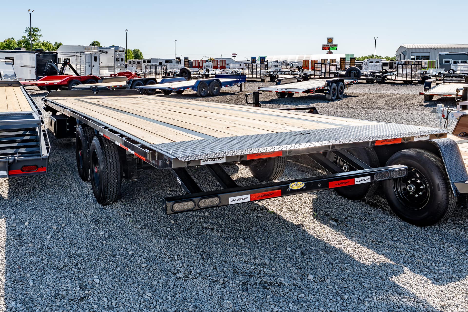 Series of flatbed trailers parked on a gravel lot at a trailer dealership, with buildings in the background and a sunny sky behind them.