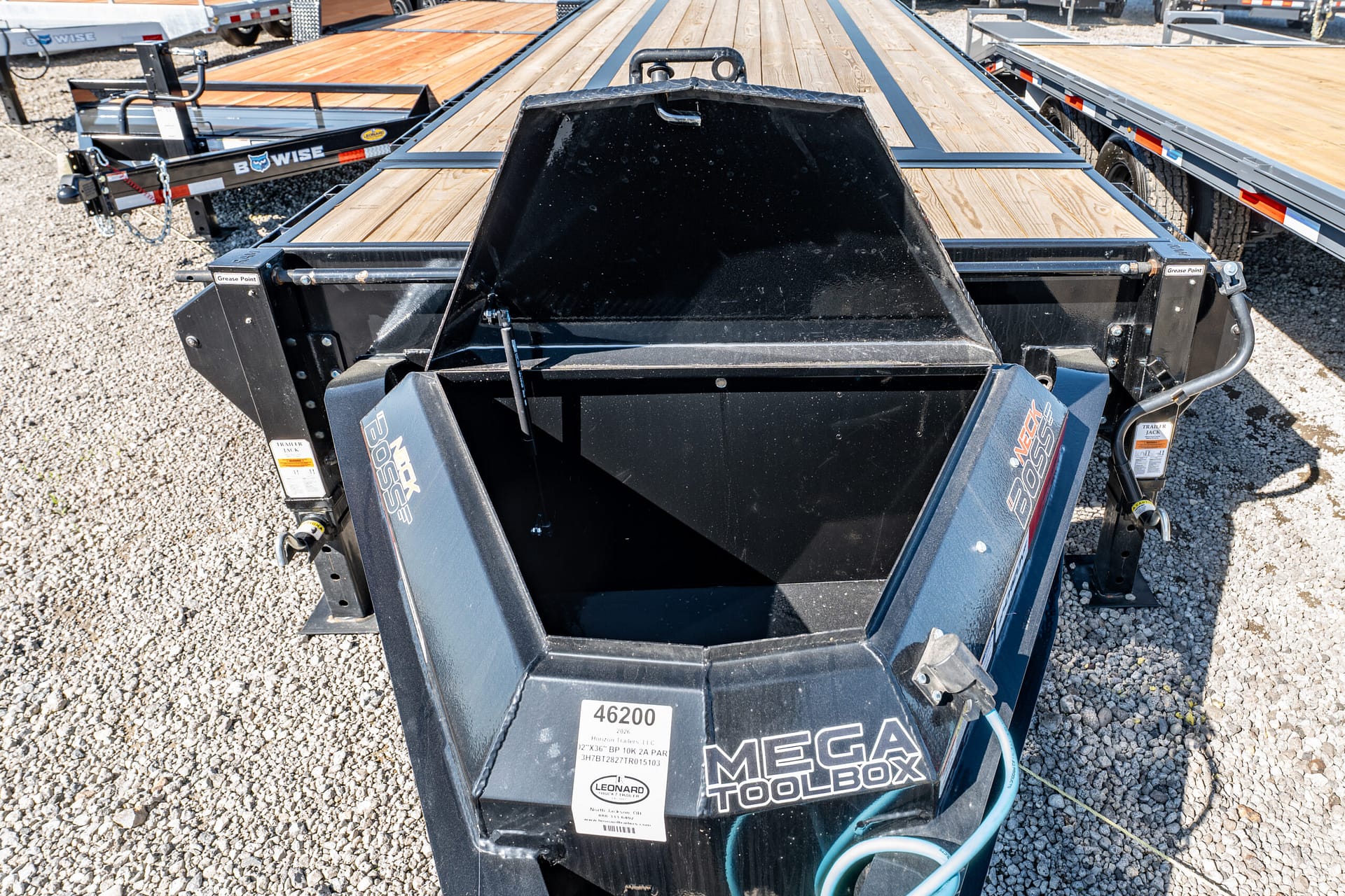 Front view of a Mega Tool Box flatbed trailer with a large black central toolbox, wooden deck planks, and gravel surface behind it.