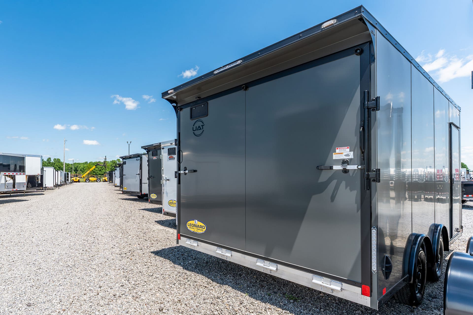 Row of gray cargo trailers parked on a gravel lot under a clear blue sky, with a large trailer in the foreground.