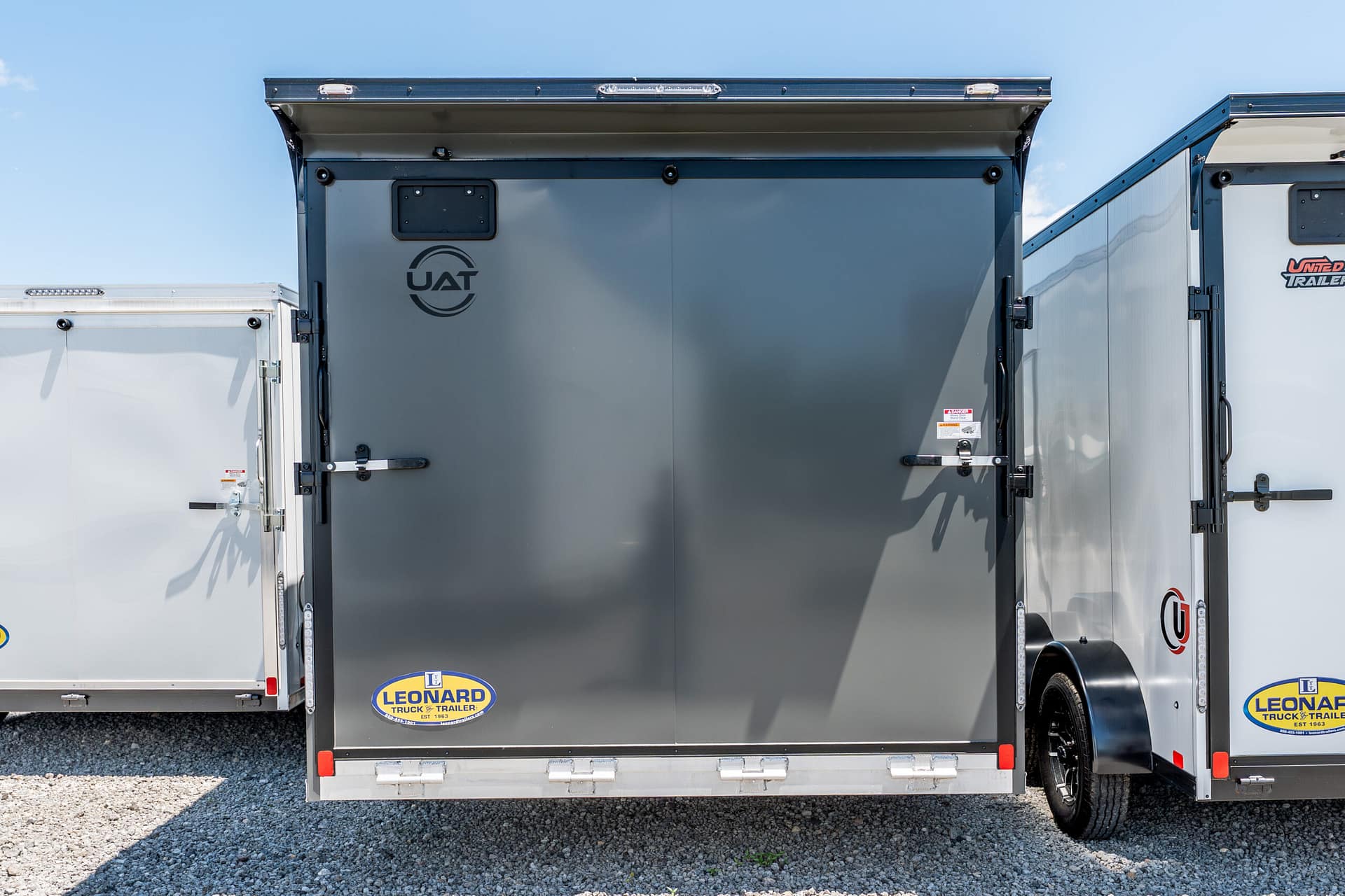 Rear view of a gray cargo trailer with closed doors; Leonard Truck & Trailer logos visible at the bottom corners, parked on gravel under a clear blue sky
