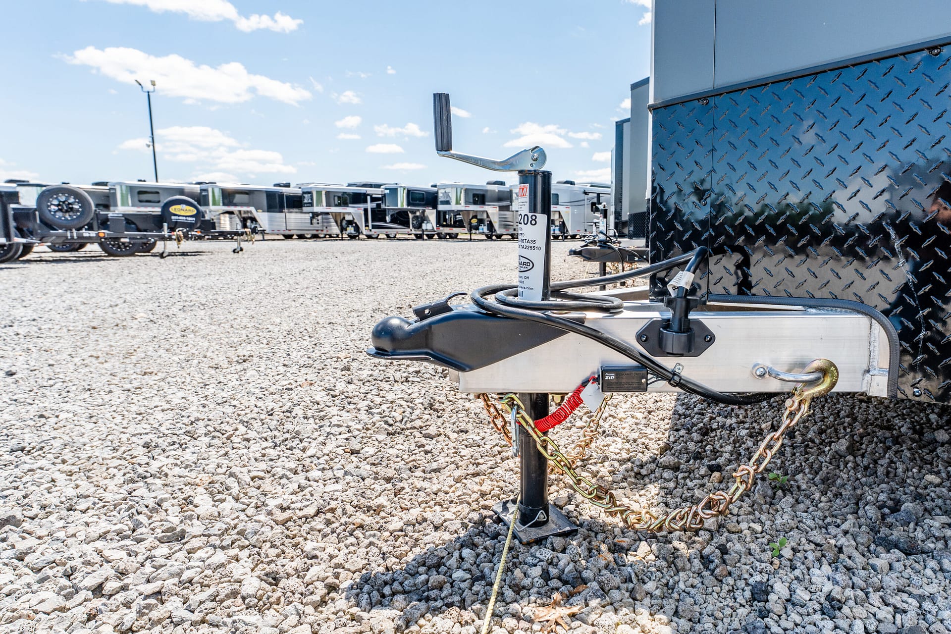 Close-up of a trailer hitch and safety chains on a gravel lot, with a diamond-plate utility box and parked campers in the background.