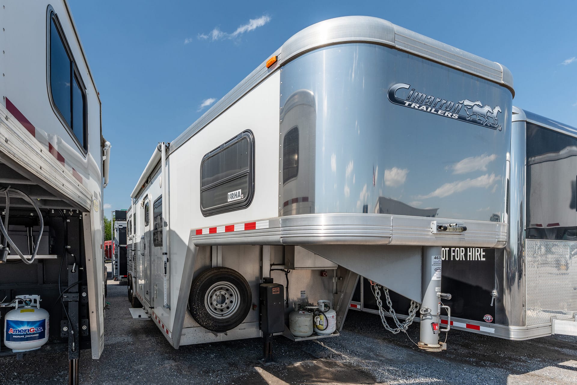 Shiny silver Cimarron trailer with logo, parked between other trailers under a blue sky.