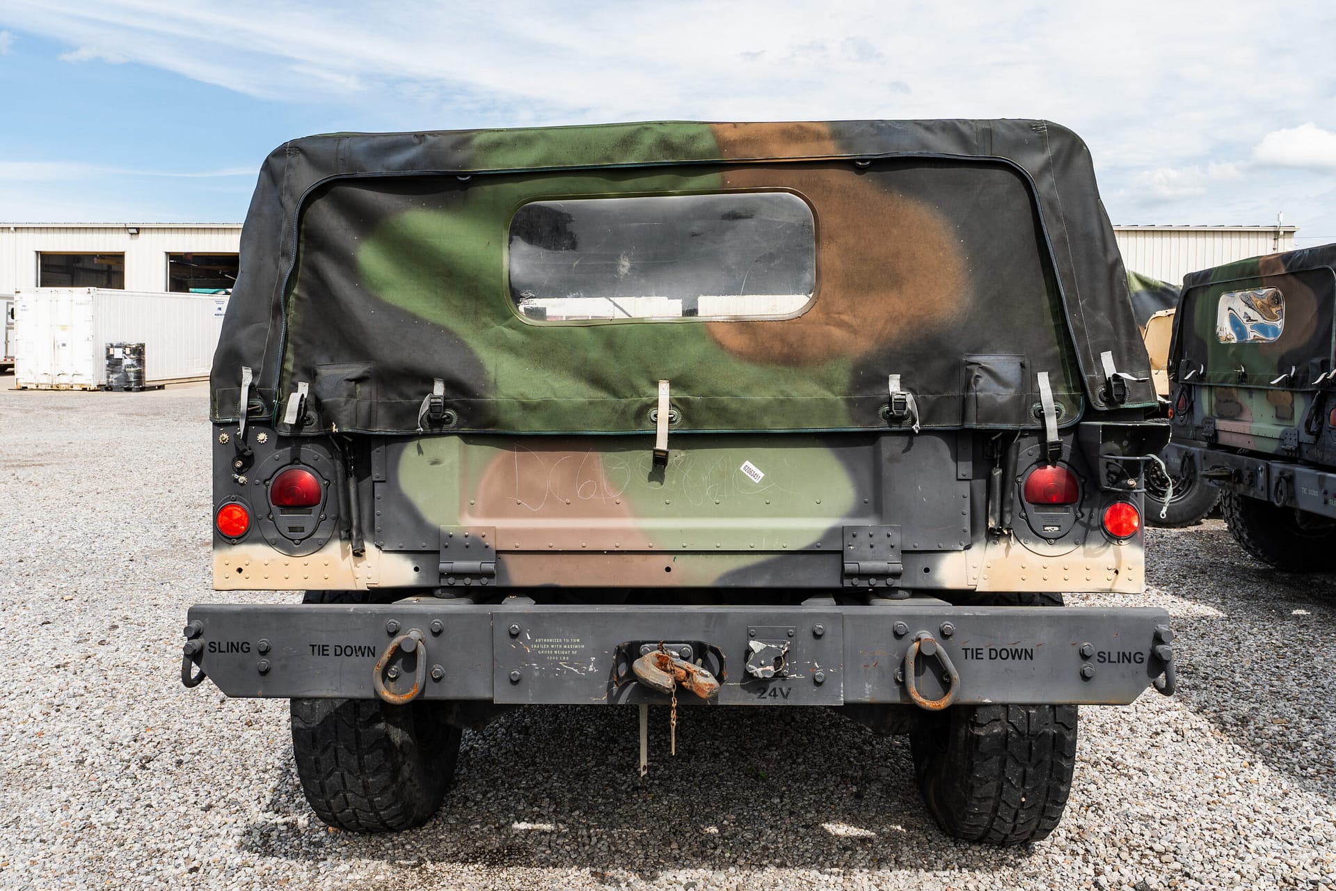 Rear view of camouflaged military trailer with canvas cover, red taillights, and tiedown hardware on a gravel lot