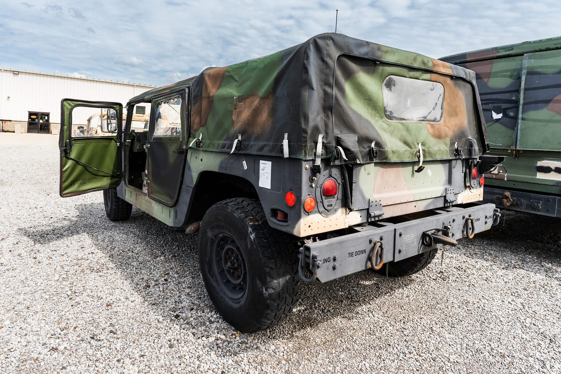 Camouflage military cargo truck with a canvas cover and an open rear door in a gravel lot.