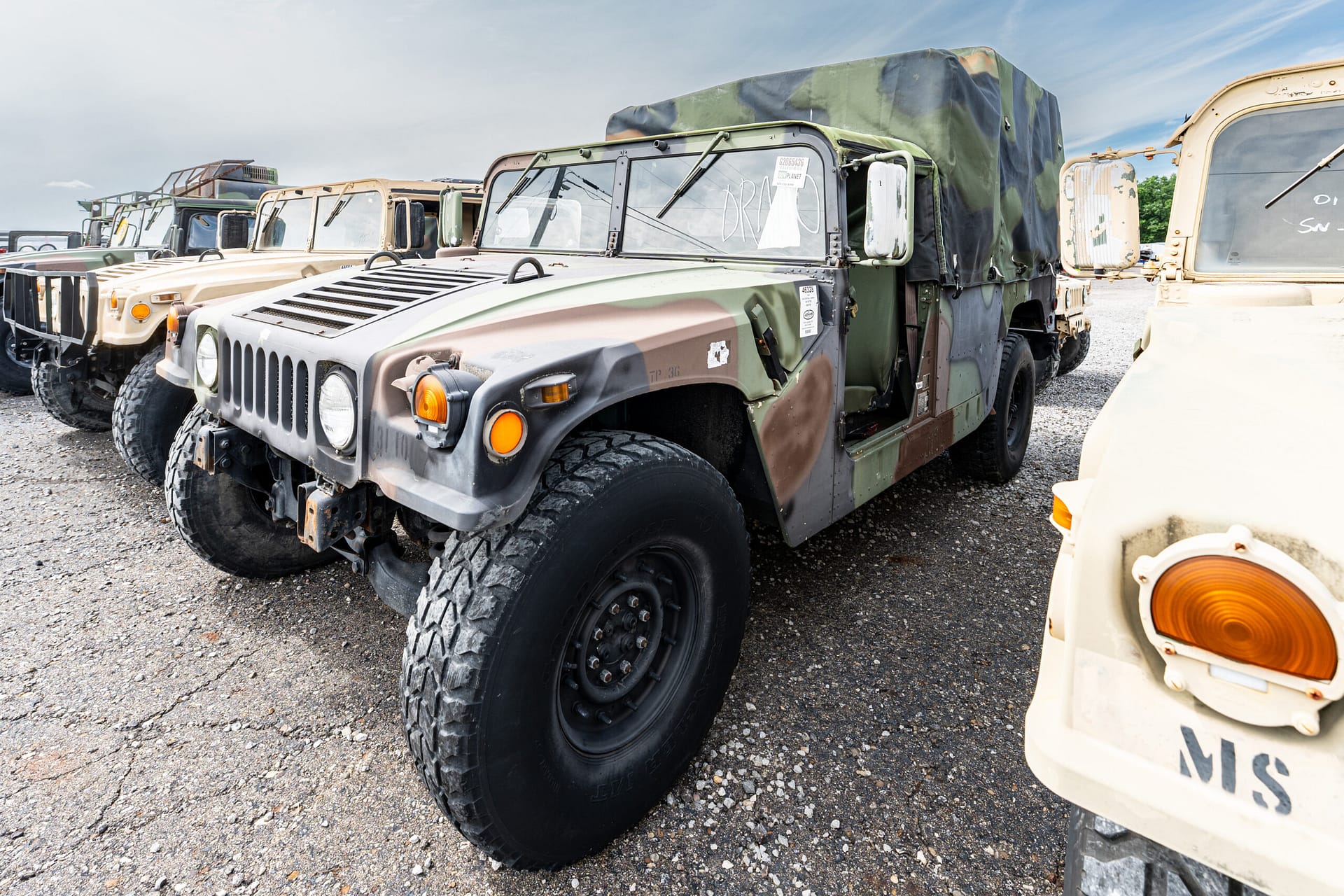 Line of camouflaged military transport trucks parked on a gravel lot, viewed from the front-left.