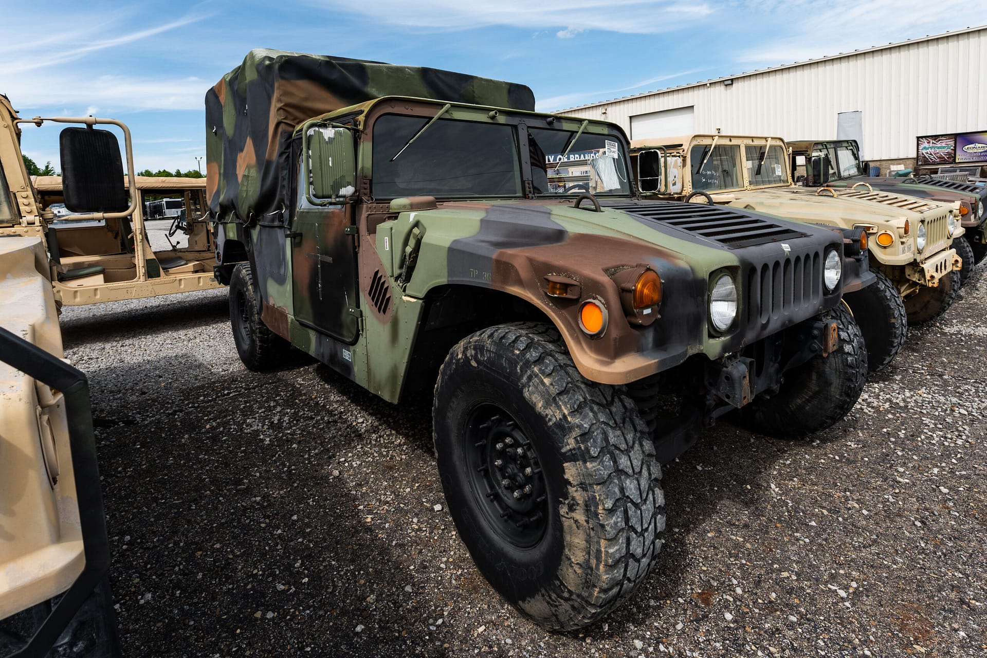 Camouflage military Humvee in a row of similar vehicles outside a light industrial building, on a gravel lot.