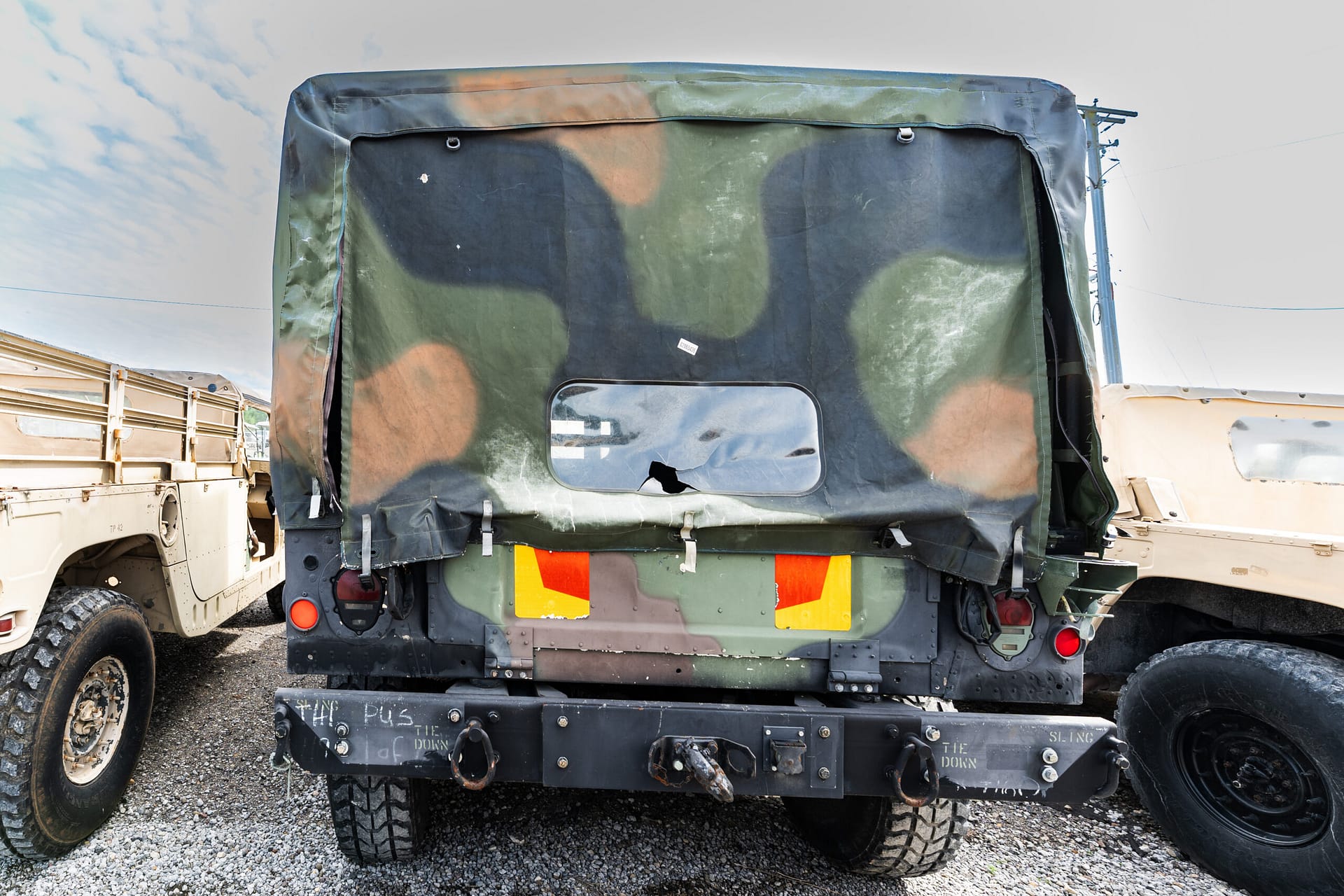 Rear view of camouflage-painted military cargo truck with tarp-covered cargo area and reflective safety panels on a gravel lot.