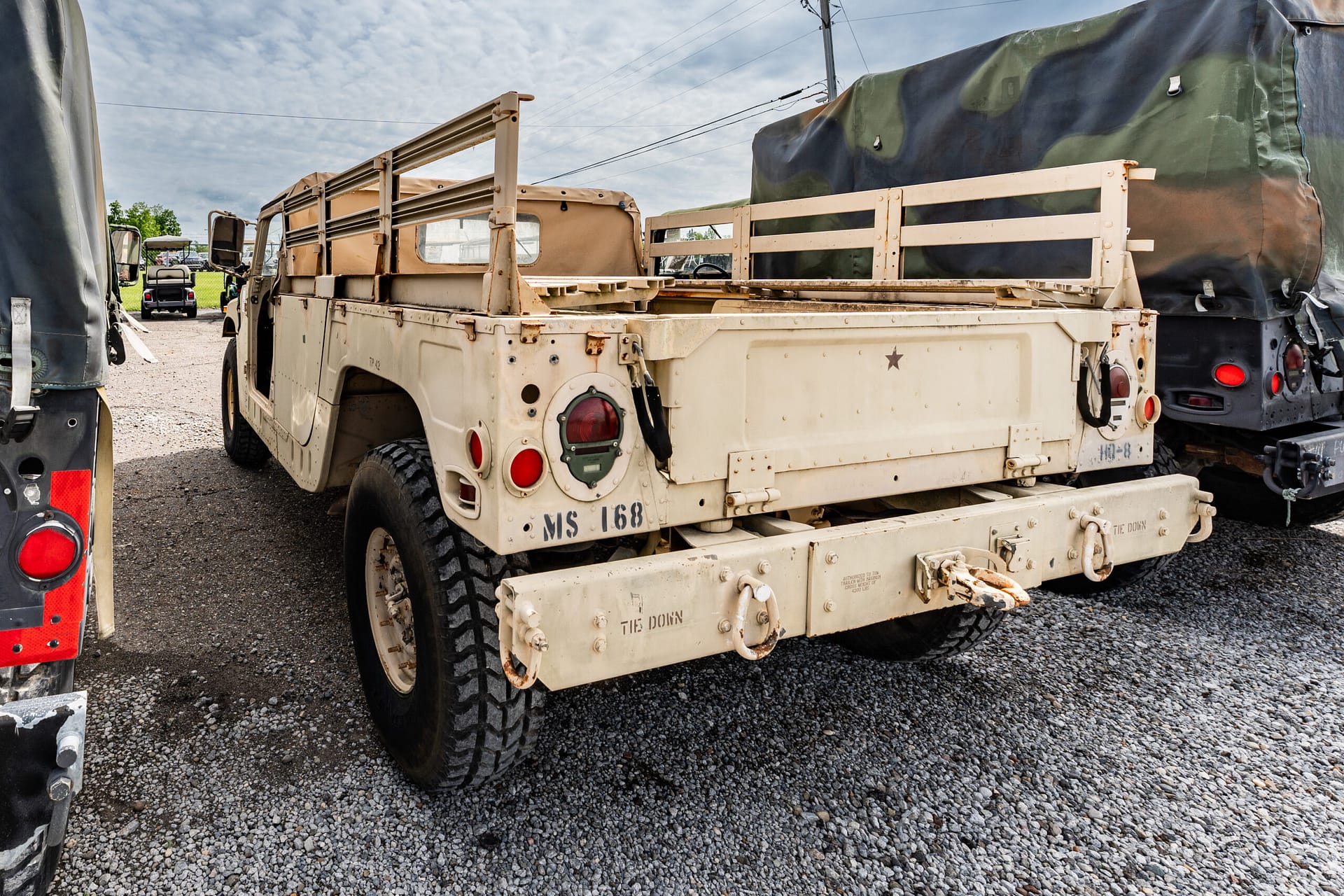 Rear view of a beige military transport truck with open cargo bed and spare wheel, parked on a gravel lot.