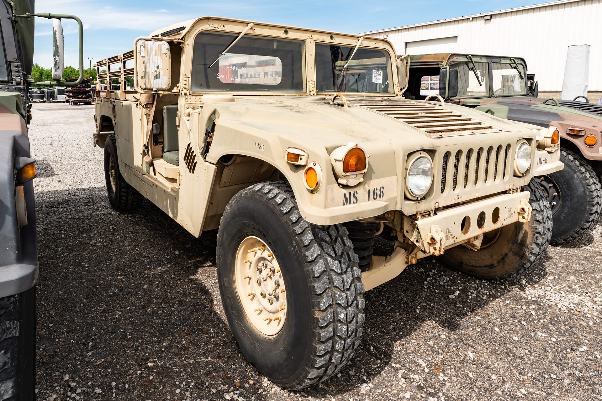 Beige military Humvee parked in a lot with others, front view showing large tires and grill badges