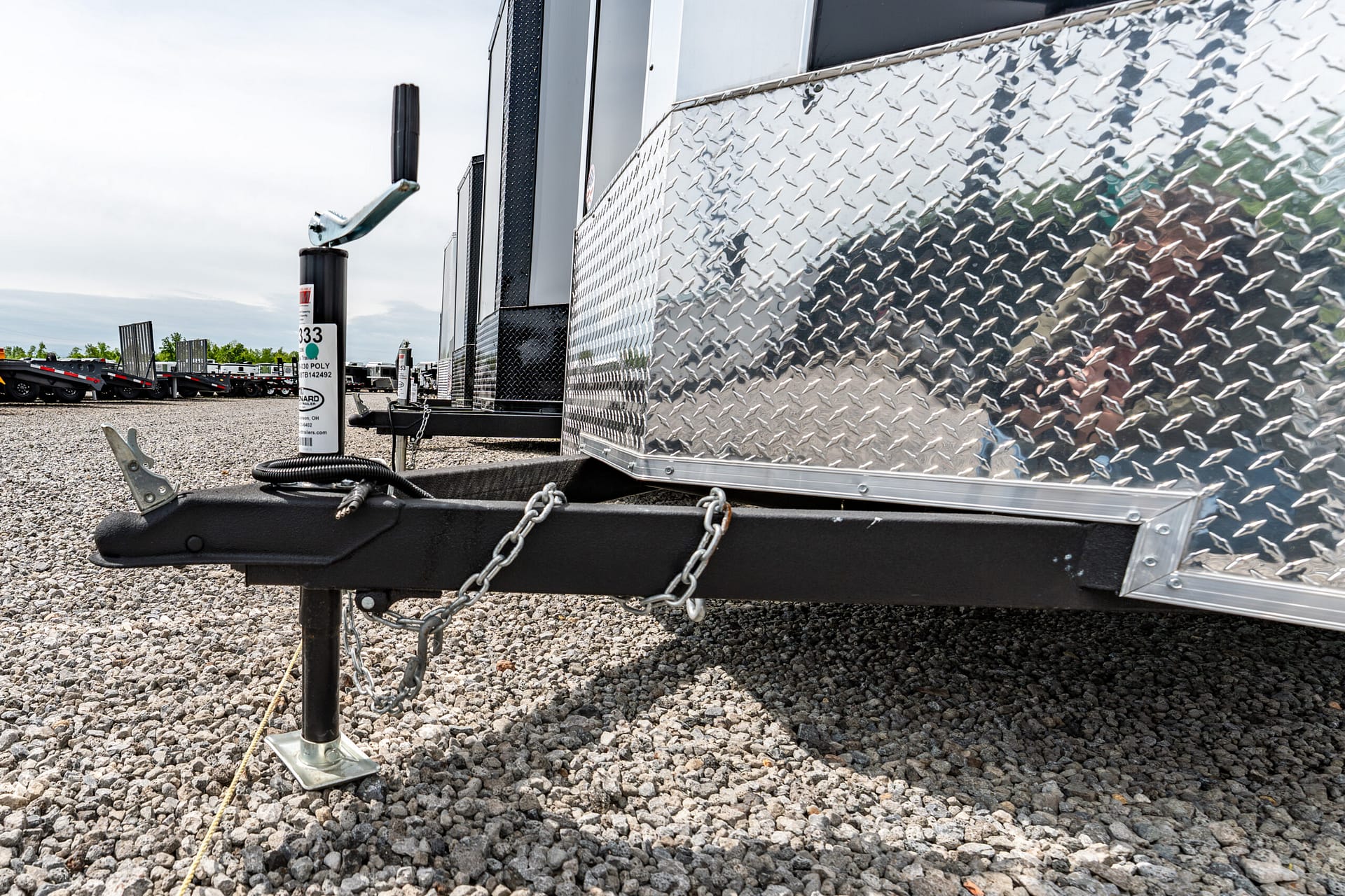 Close-up of a trailer hitch and the diamond-plate side of a utility trailer on a gravel lot, with safety chains visible.