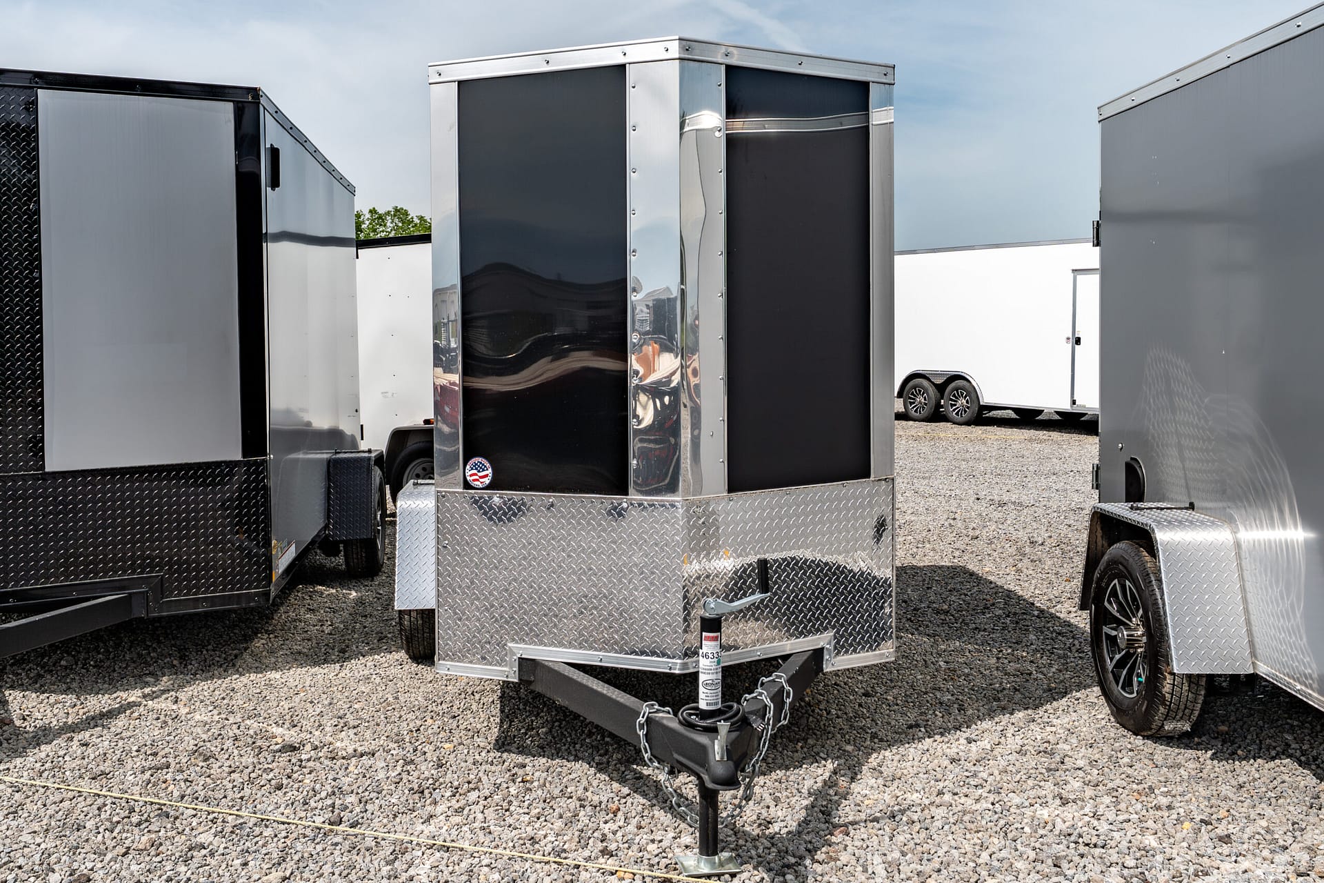 Row of enclosed cargo trailers with diamond-plate fronts parked on gravel under a blue sky; central trailer has black panels and chrome trim, hitch visible in foreground