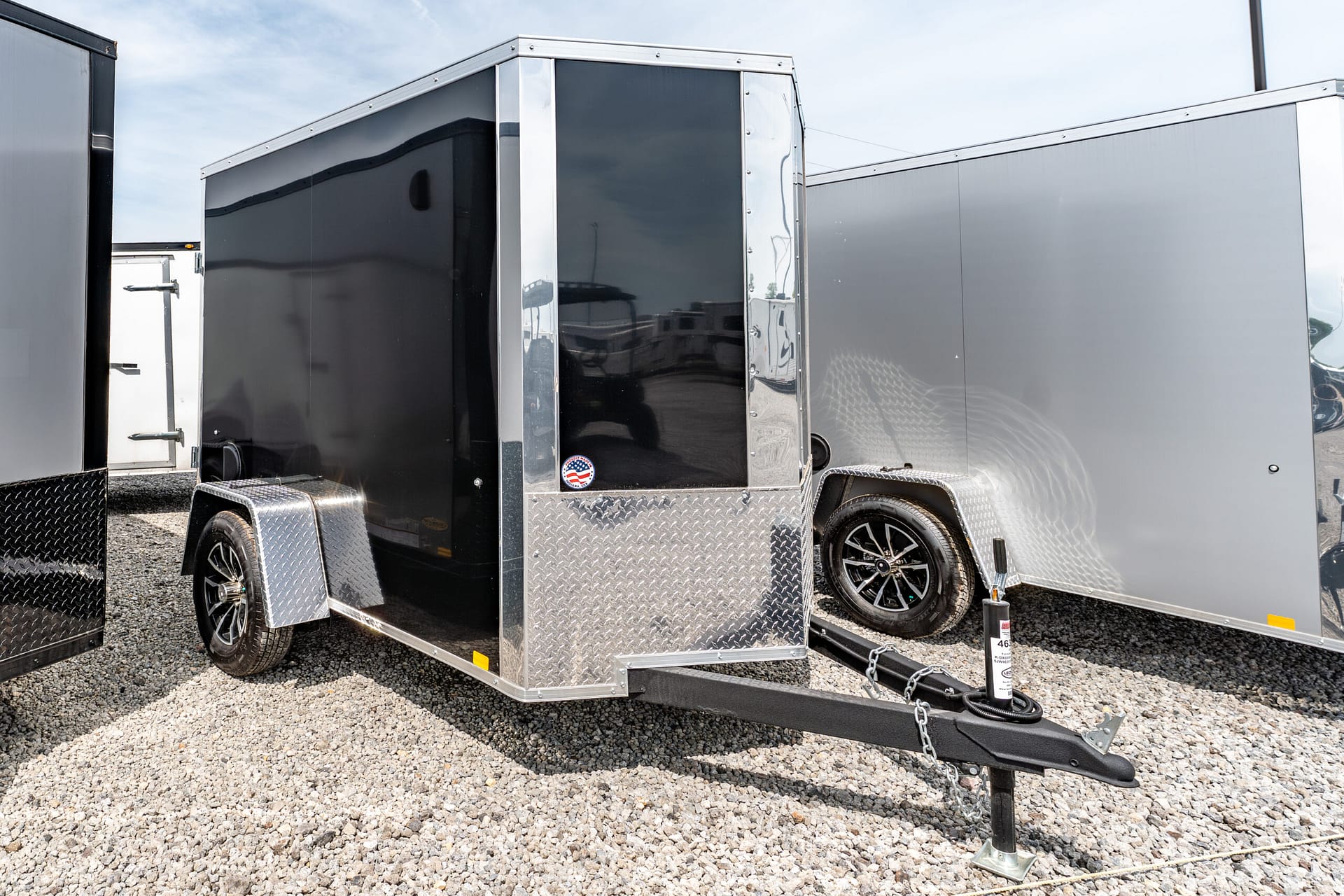 Black enclosed trailer with chrome trim and diamond-plate front, parked on gravel, hitch and wheel visible