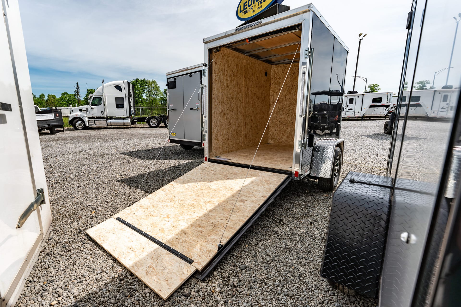 Open cargo trailer with plywood floor and lowered ramp, parked in a gravel lot with other trucks nearby.