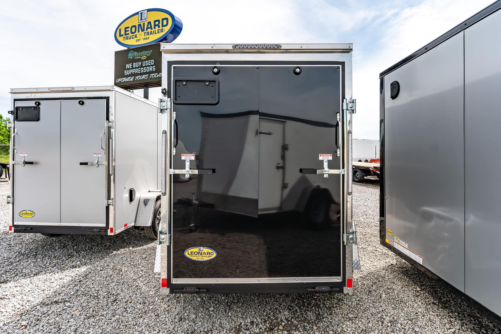 Open black trailer rear between two silver trailers with Leonard Truck & Trailer logos visible on lower corners, on a gravel lot with a blue sky.
