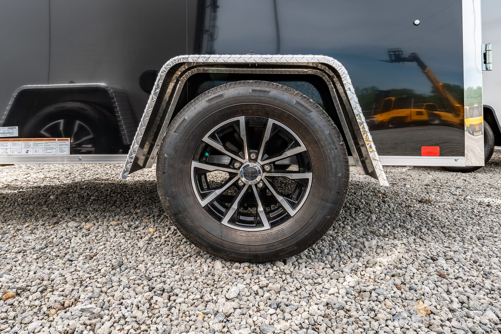 Close-up of a trailer wheel with a diamond-plate fender on a black trailer, gravel ground beneath.