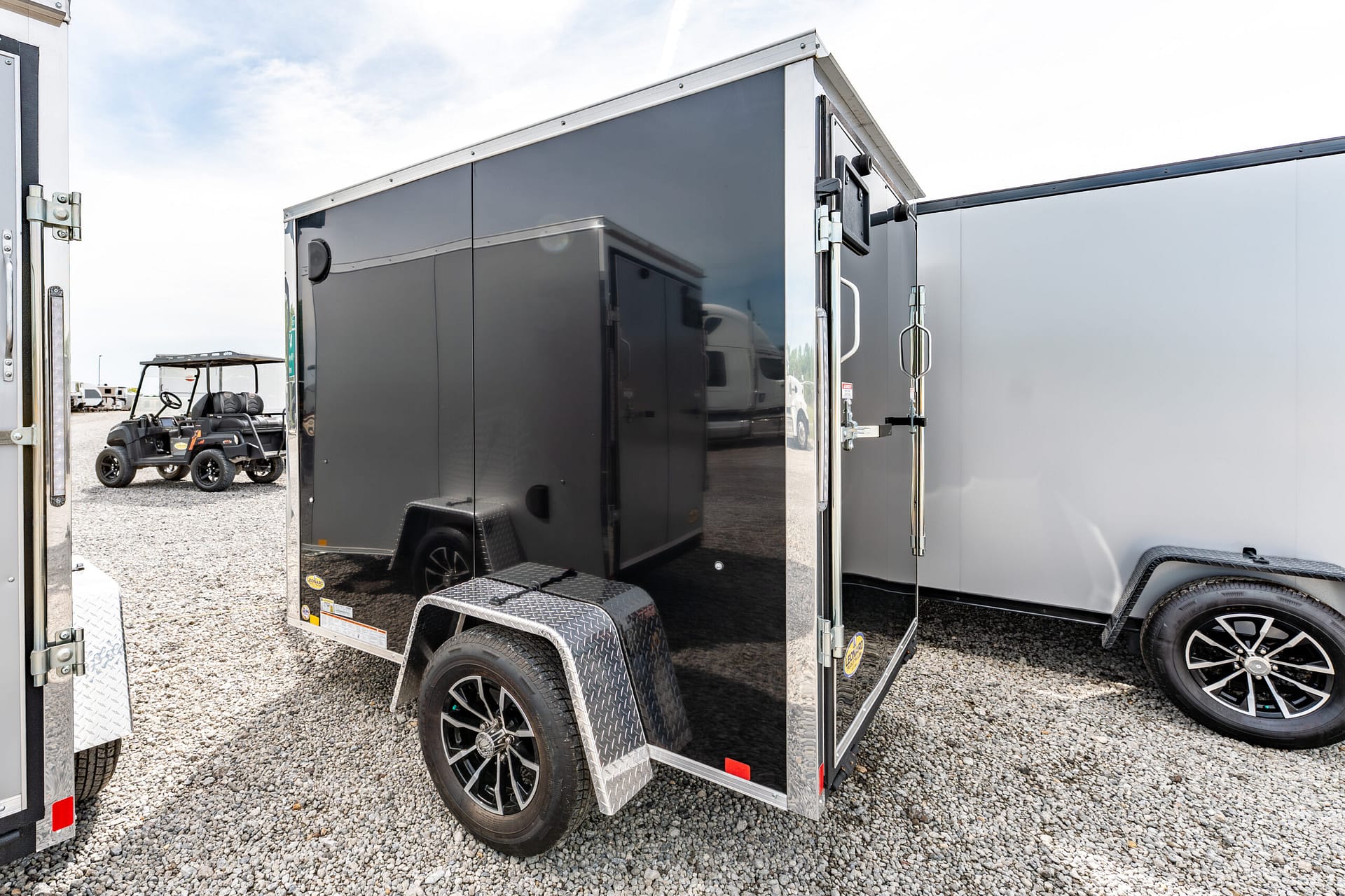 Dark gray enclosed cargo trailer with metal diamond-plate steps and black wheels, parked on a gravel lot.