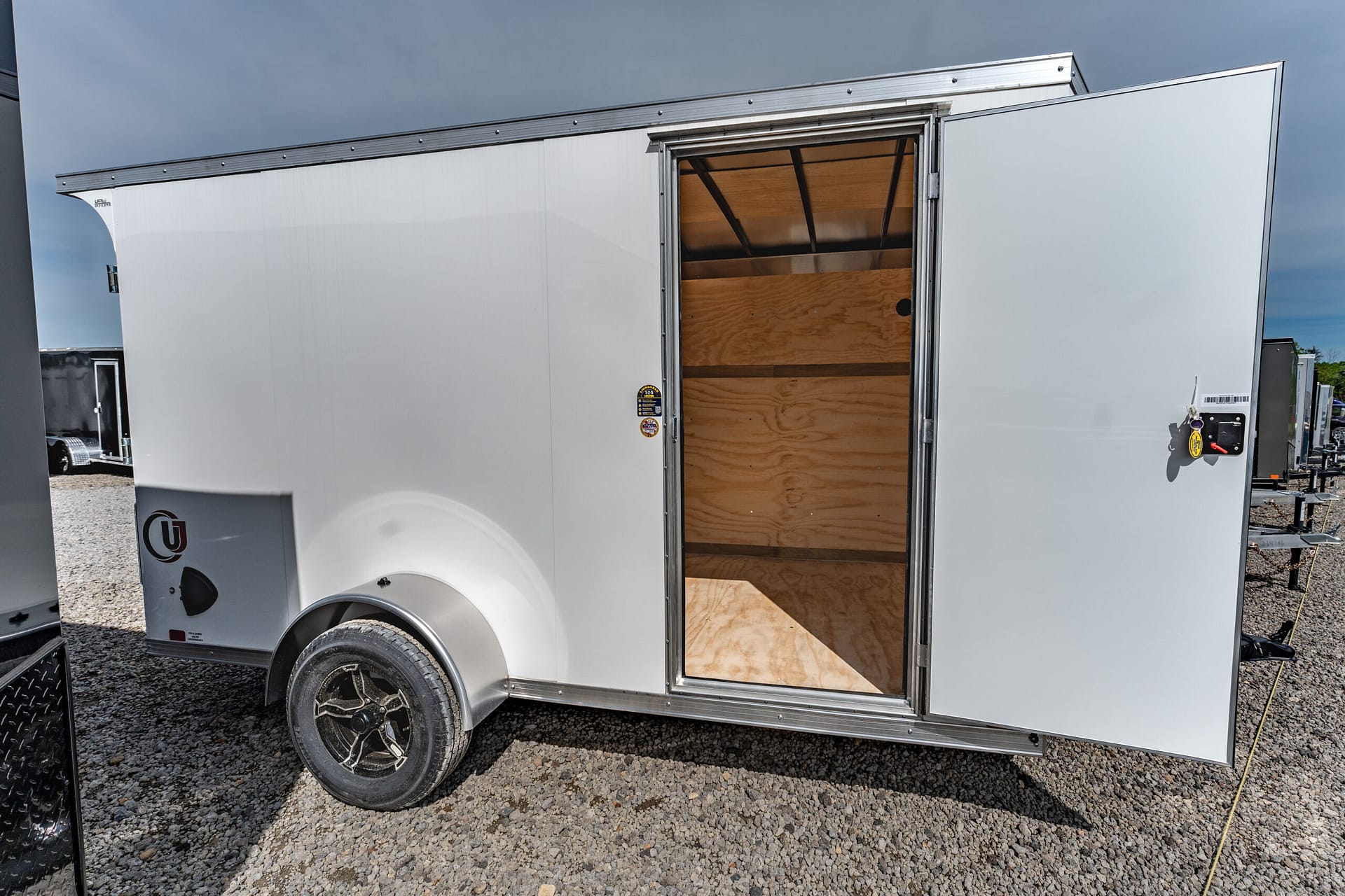 White enclosed trailer with its back door open, exposing wooden interior shelves and plywood walls on a gravel lot