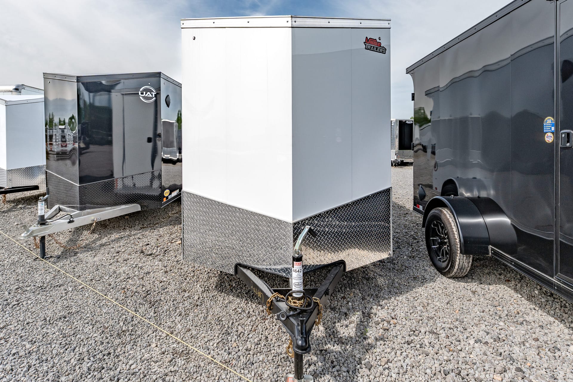 Row of enclosed cargo trailers on a gravel lot; white center trailer flanked by black and gray trailers.