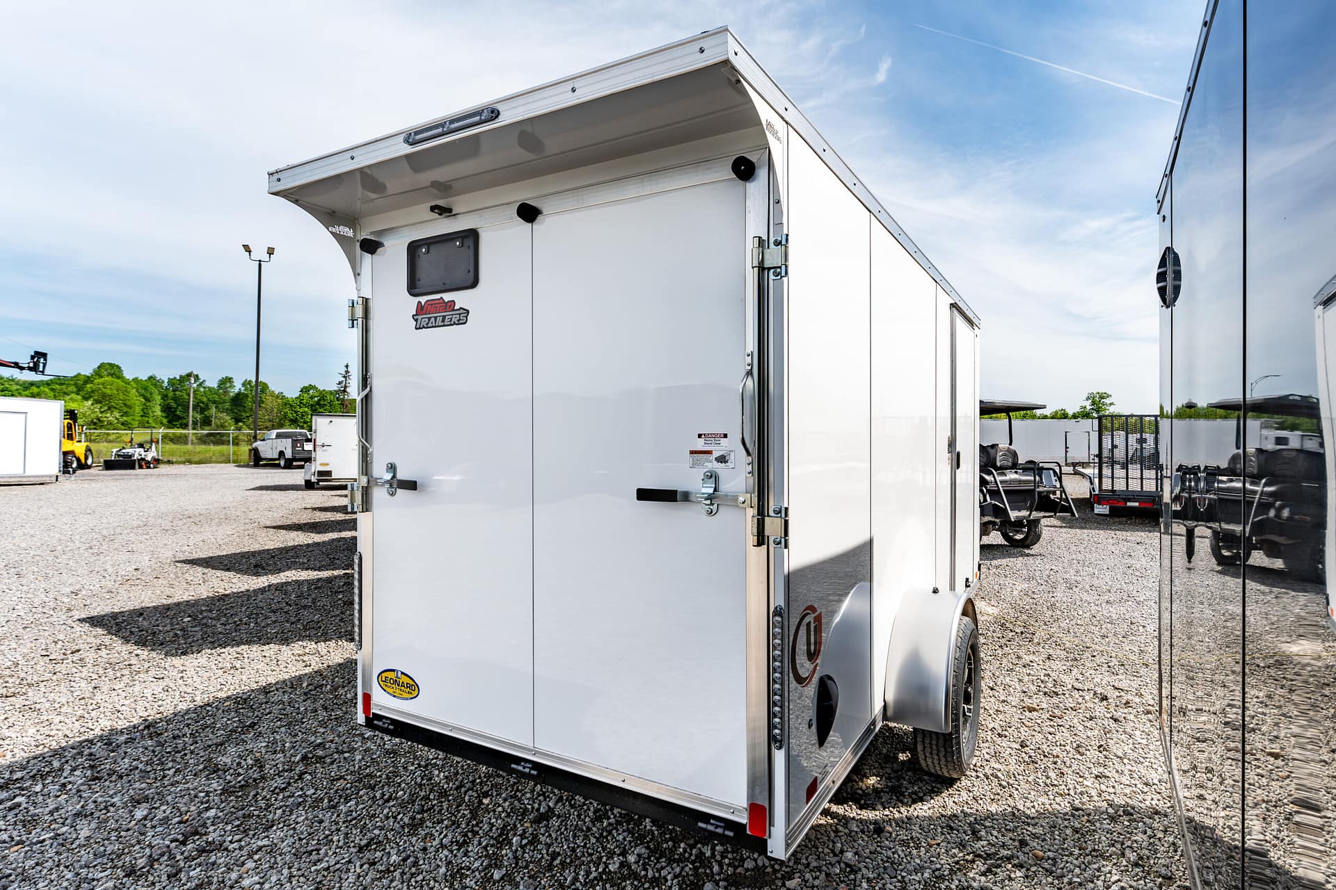 Rear view of a white enclosed trailer on a gravel lot, with other trailers in the background, under a blue sky.