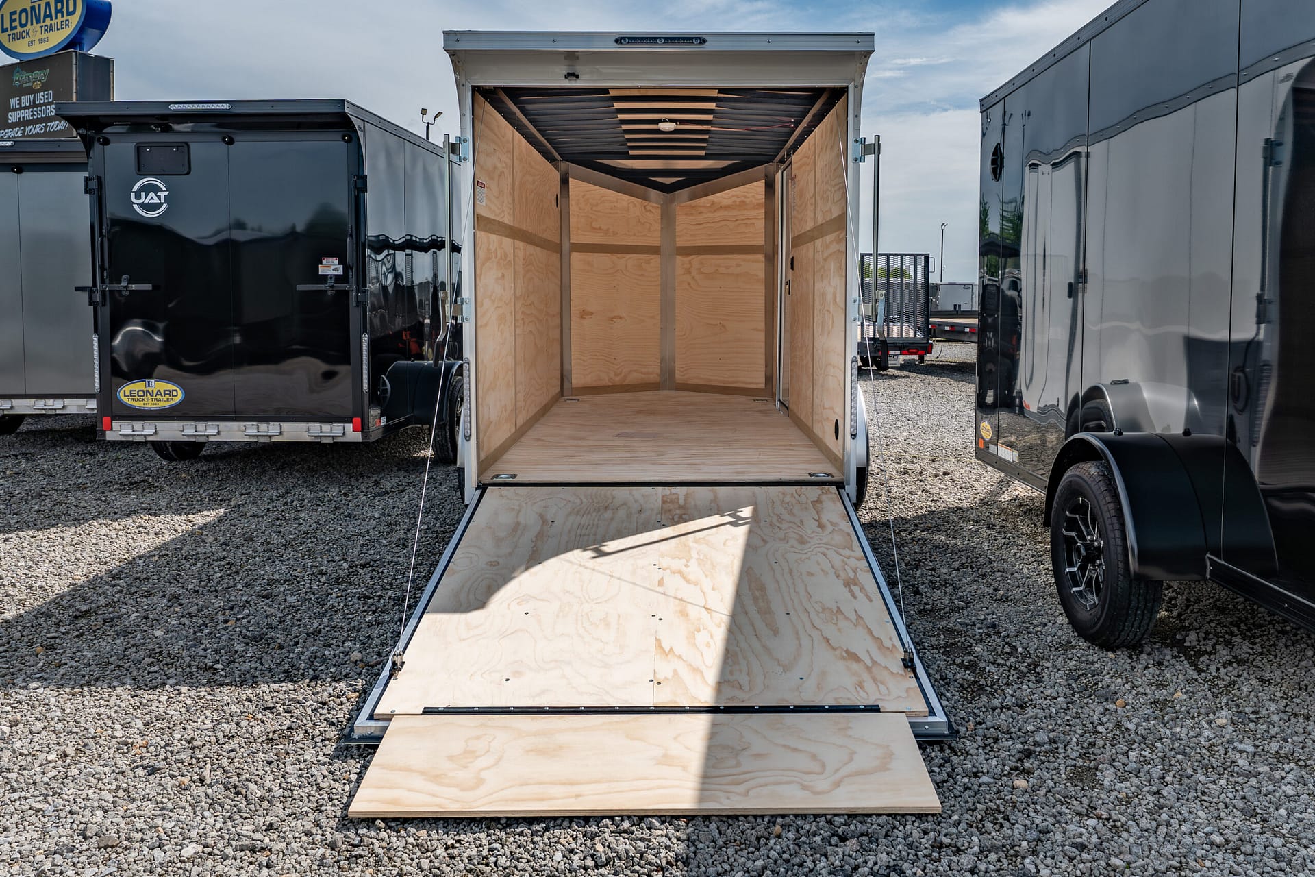 Open cargo trailer with plywood walls and ramp lowered to gravel, parked between black and gray trailers on a gravel lot.