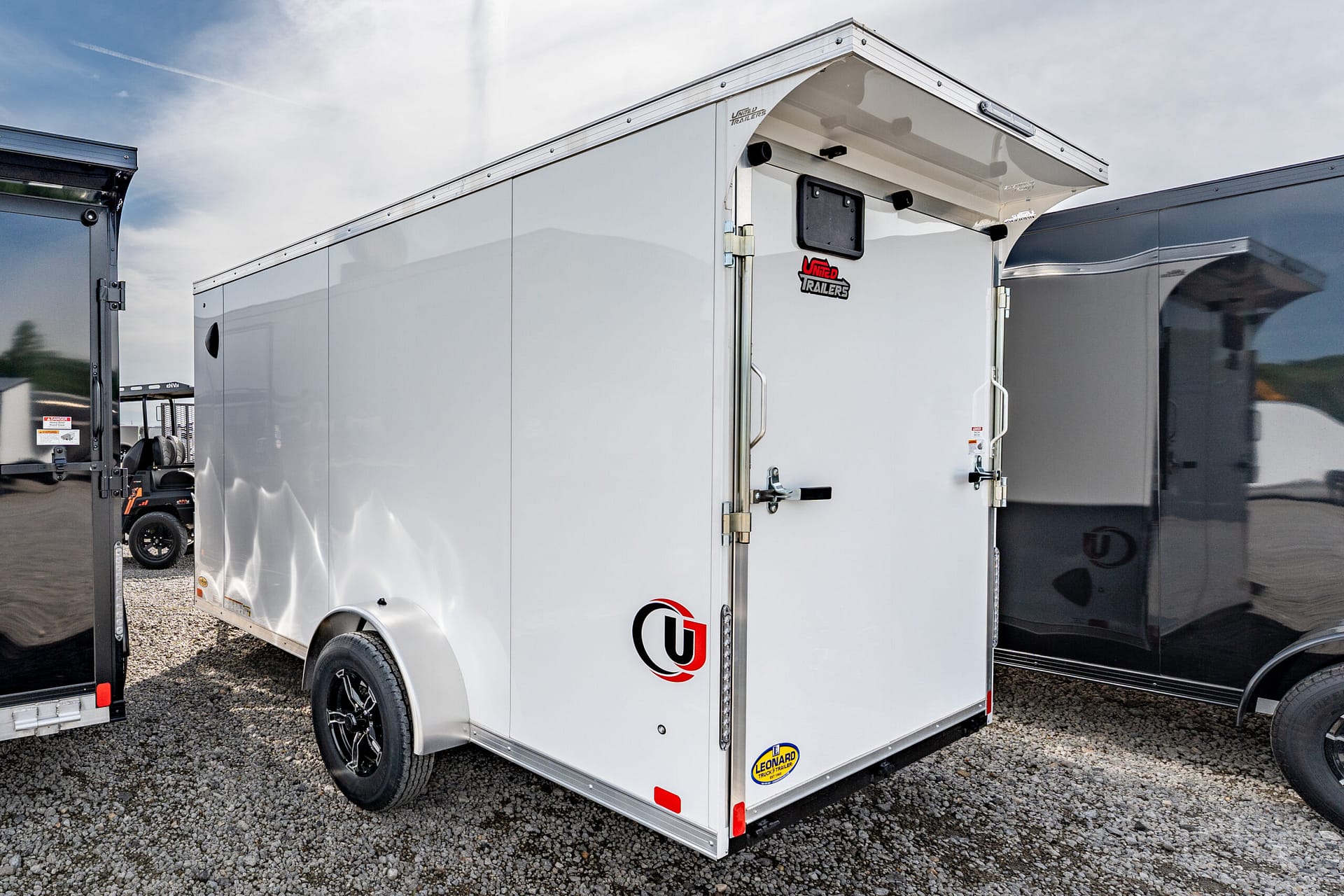 White enclosed cargo trailer with dual rear doors parked on a gravel lot, with another trailer nearby.