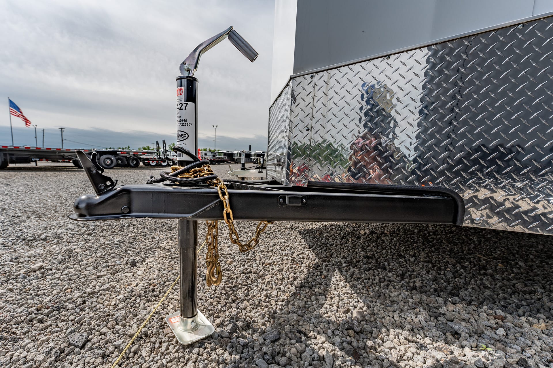 Close-up of a trailer hitch and its jack with safety chains on a gravel lot, next to a diamond-plate trailer side and out-of-focus American flag in the background.