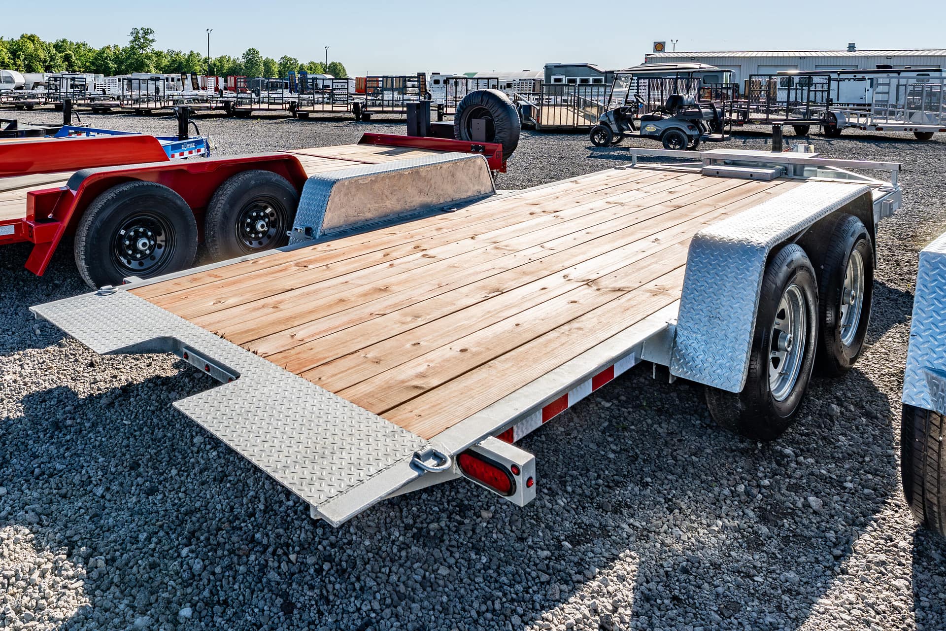 Flatbed trailer in a gravel lot with other trailers and equipment in the background, under a clear blue sky.