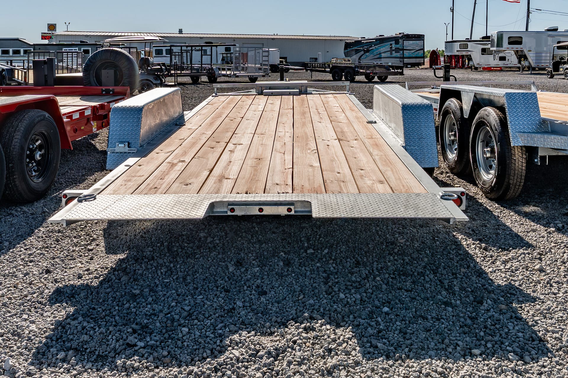 Flatbed trailer with a wooden deck and metal fenders on a gravel lot, with other trailers and RVs in the background.