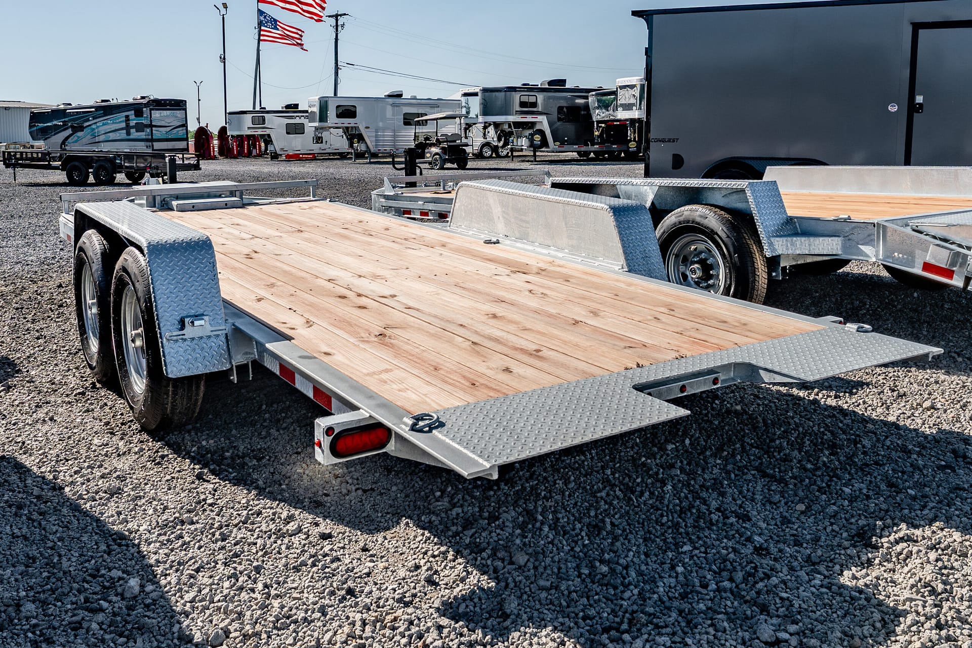 Dual-axle flatbed trailer with a wooden deck and diamond-plate fenders in a gravel RV lot.