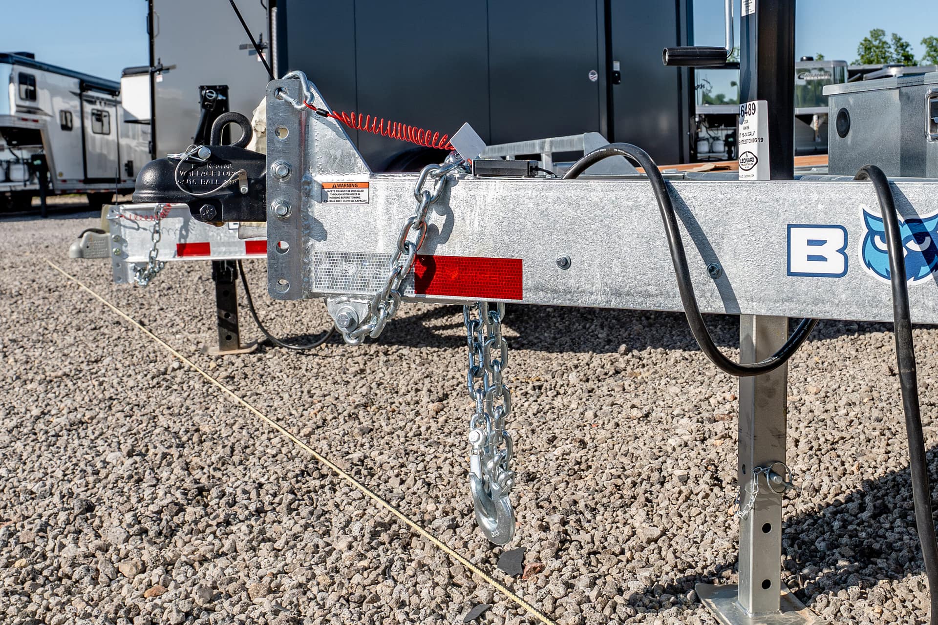 Close-up of a trailer hitch with safety chains and a red coiled cable on a gravel lot, showing a galvanized frame and hardware.