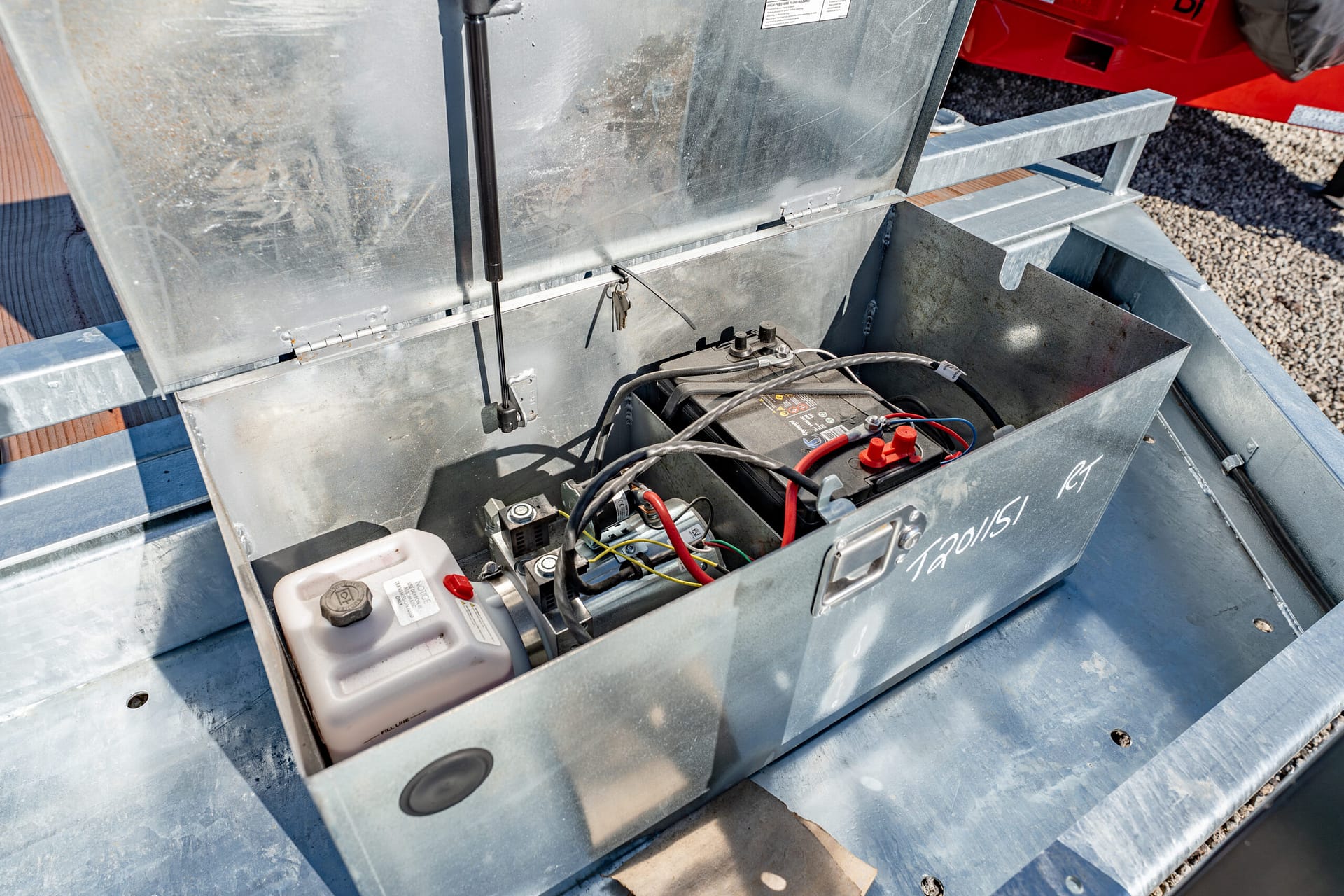 Open metal battery box on a utility trailer with a car battery and tangled cables inside the compartment.