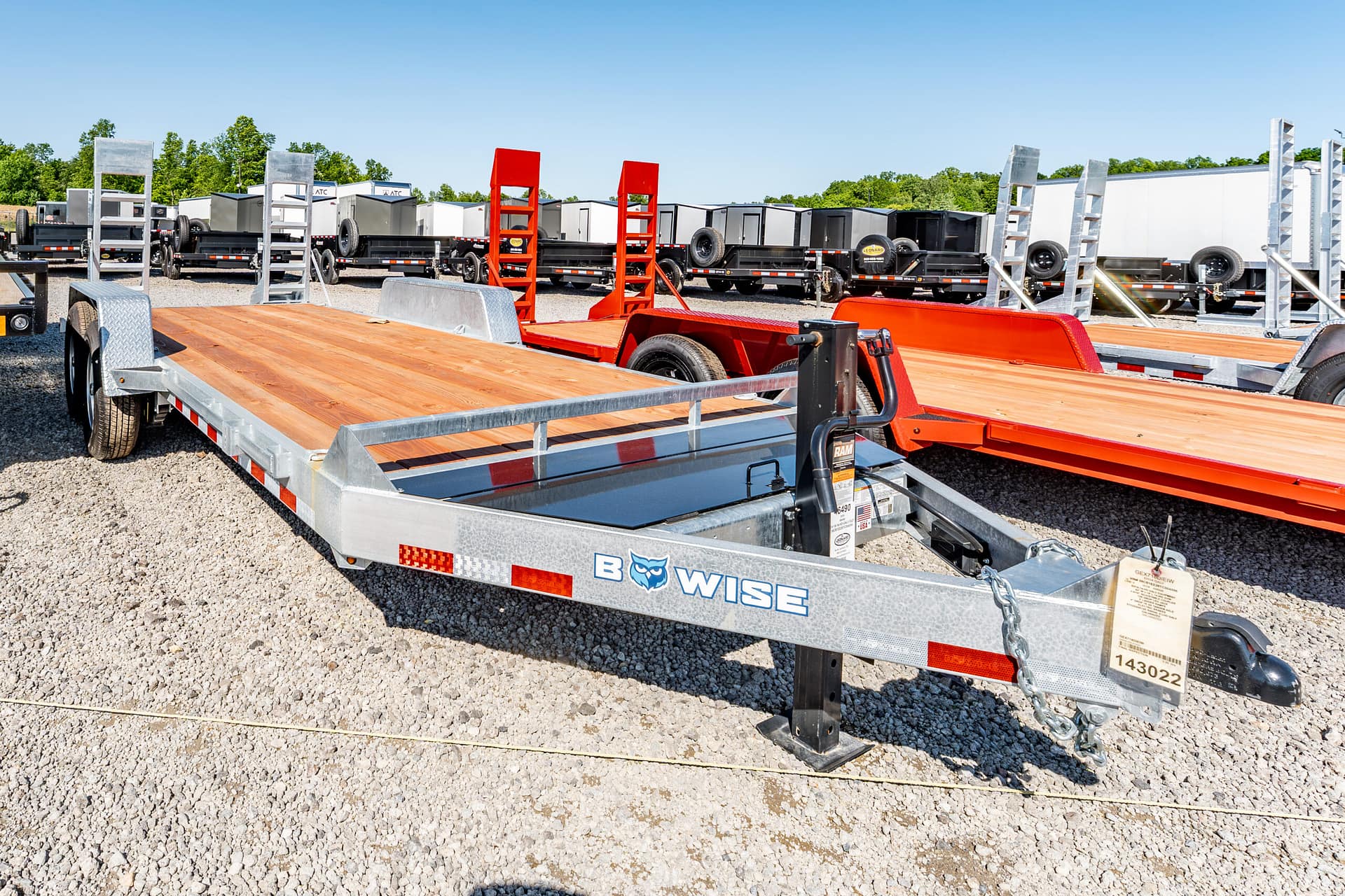 Row of flatbed trailers and orange loading ramps in a gravel trailer yard under a clear blue sky.