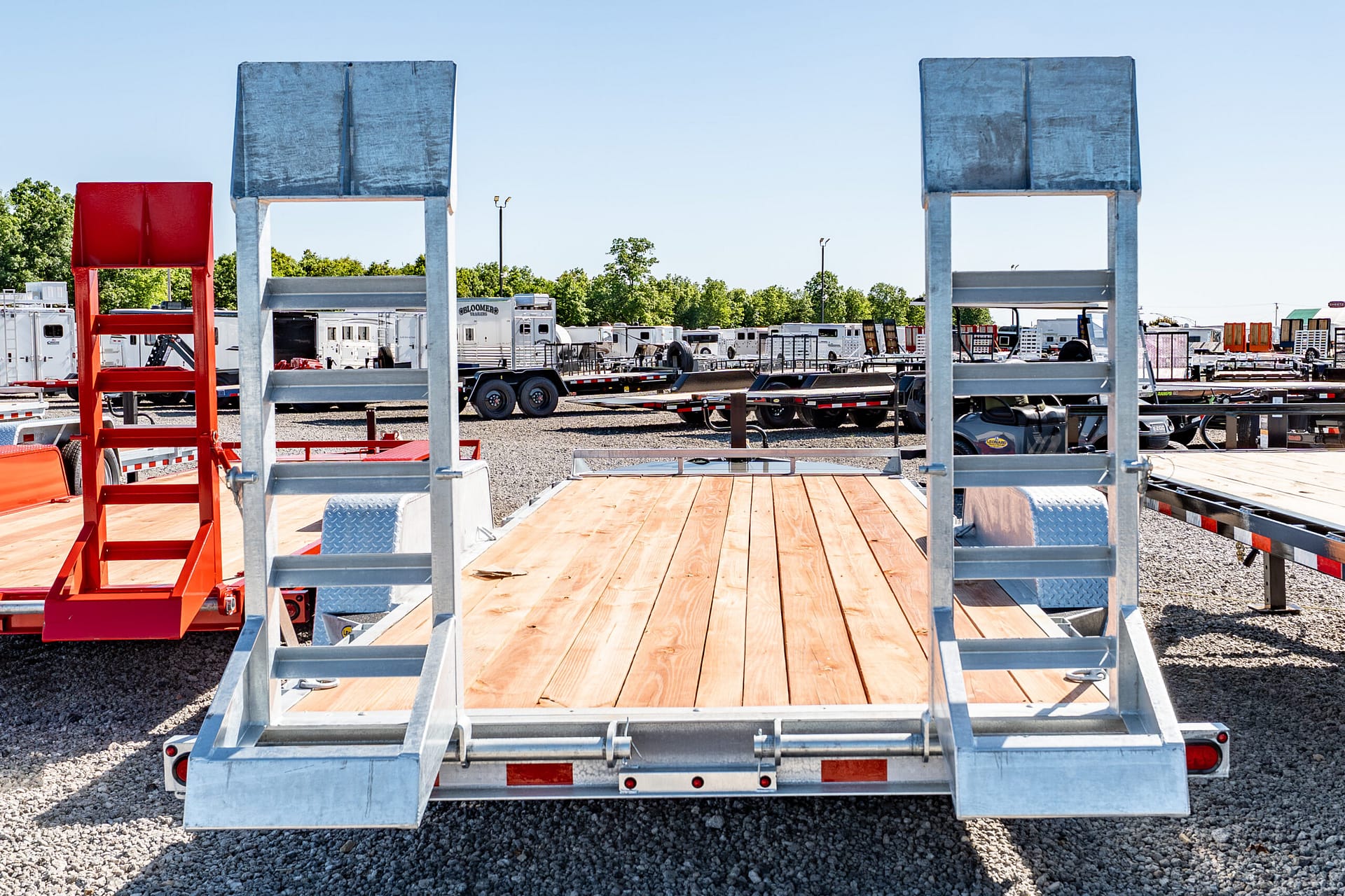 Rear view of a flatbed trailer with tall gray metal frames and a red ladder on the left, in a gravel lot with other trailers in the background.