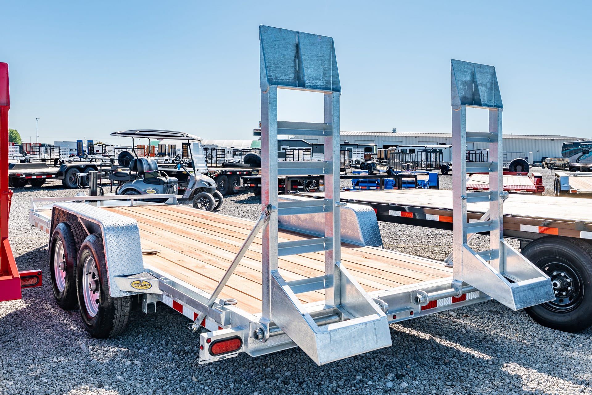 Metal-framed flatbed trailer on a gravel lot, with other trailers and equipment in the background under a blue sky.