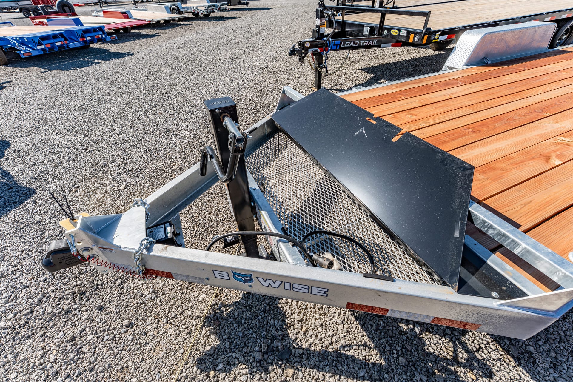 Close-up of a utility trailer hitch with safety chains on a gravel lot beside a wooden-deck trailer edge.