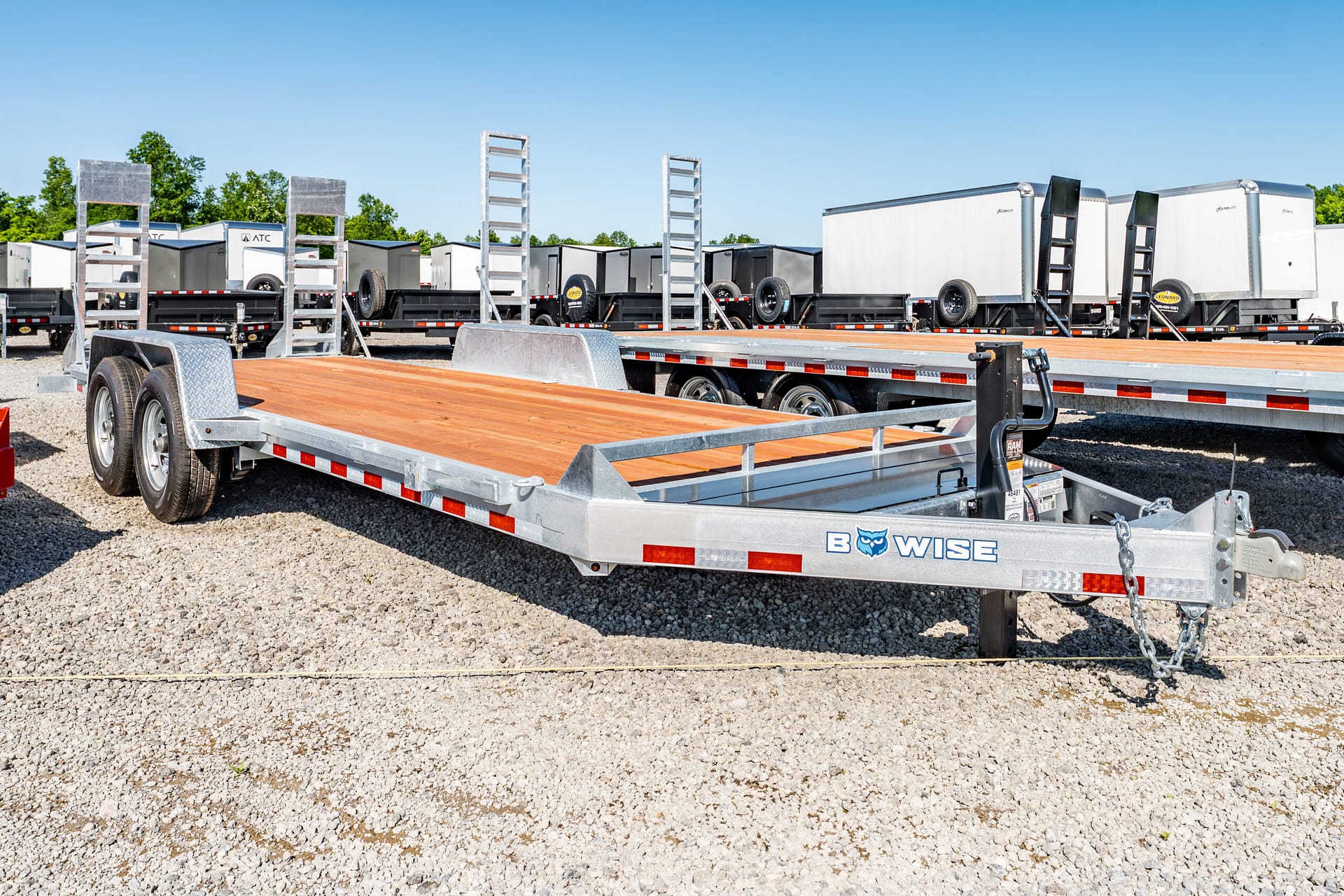 Row of aluminum flatbed trailers with wooden decks and metal ladders in a gravel lot under a clear blue sky.